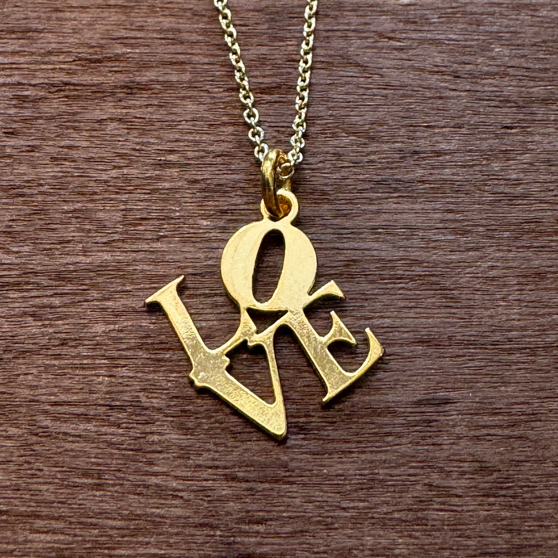 A gold-plated LOVE Necklace from GeoMetricGem lies on a wooden surface, showcasing its intricate design.