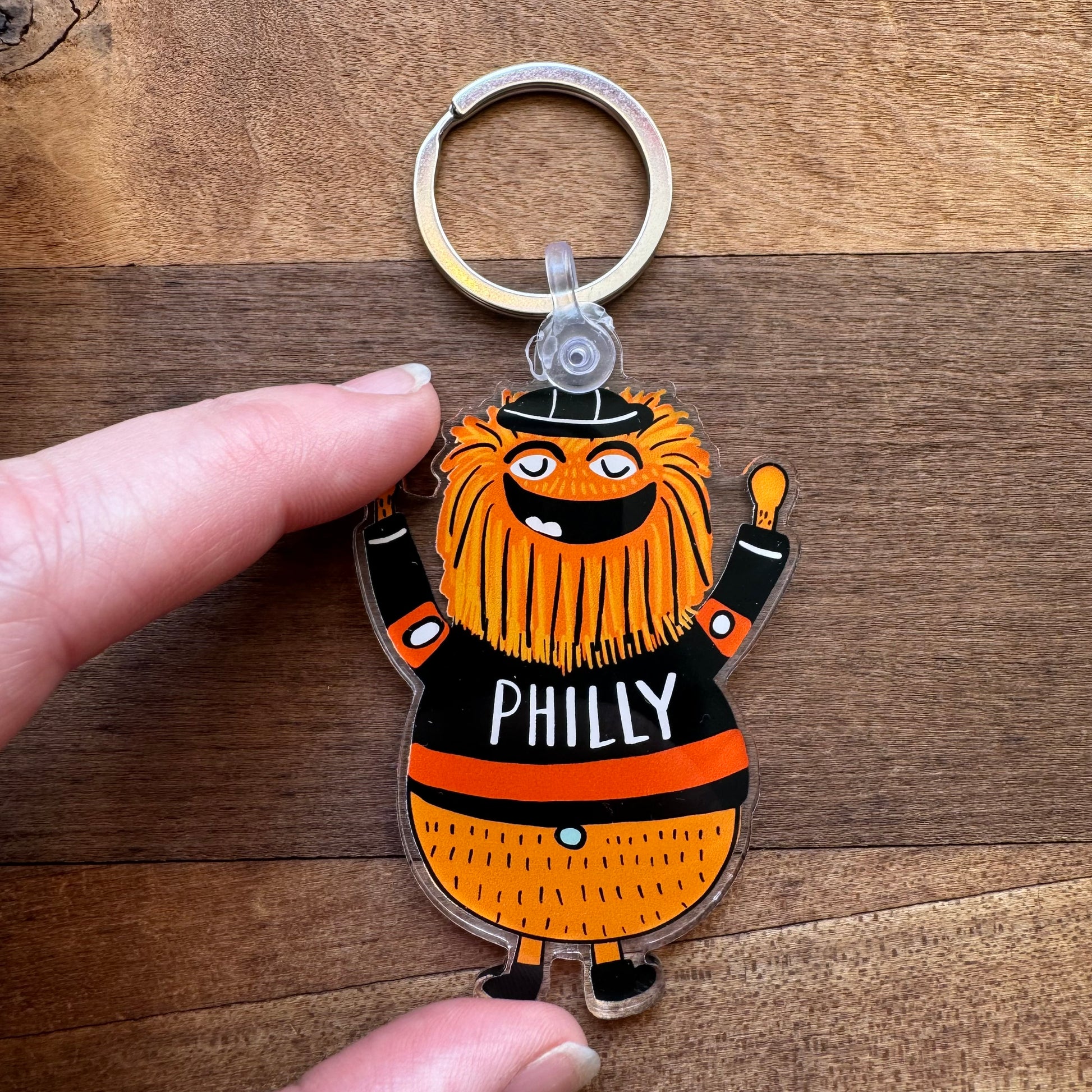 Close-up of a hand holding an Ana Thorne Gritty & Phanatic keychain, showcasing the orange-furred, smiling mascot wearing a black jersey with "PHILLY" written on it. The mascot stands on a wooden surface.