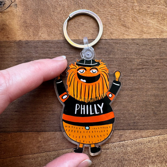 Close-up of a hand holding an Ana Thorne Gritty & Phanatic keychain, showcasing the orange-furred, smiling mascot wearing a black jersey with 