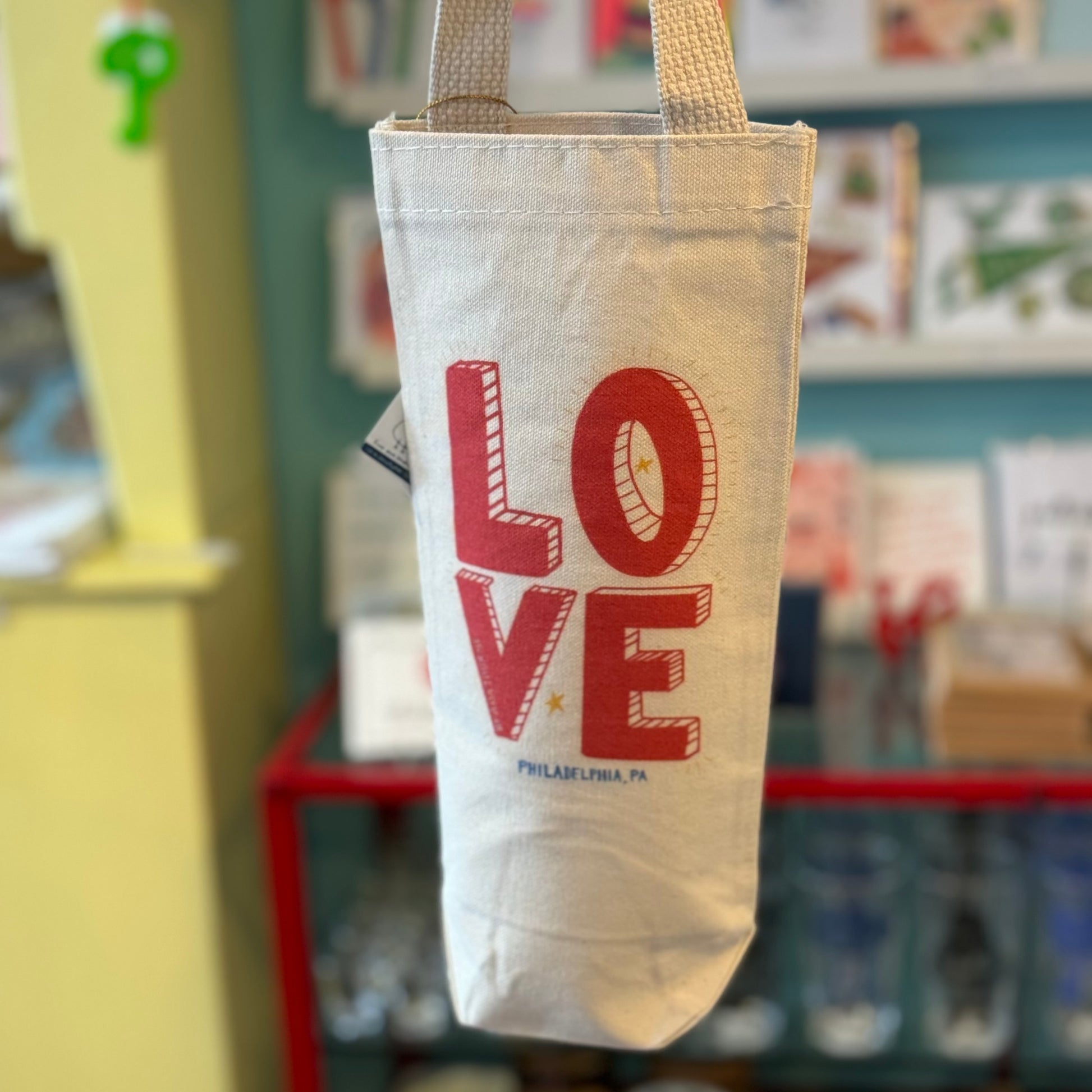 A canvas tote bag with the word "LOVE" printed on it is hanging on display in a store. The text includes "Philadelphia, PA" underneath. In the background, various other products are visible, along with a stylish LOVE Wine Tote by Ana Thorne to match your needs.