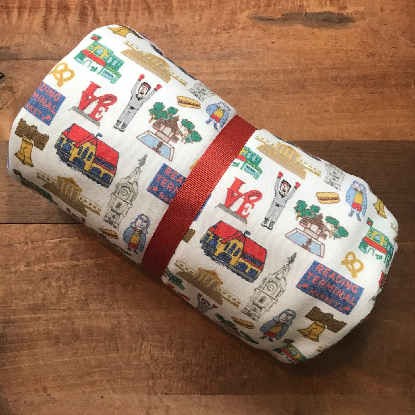 A Philly Baby Blanket by Ana Thorne with various iconic symbols and landmarks printed on its organic cotton fabric, secured with a red band, displayed on a wooden surface.