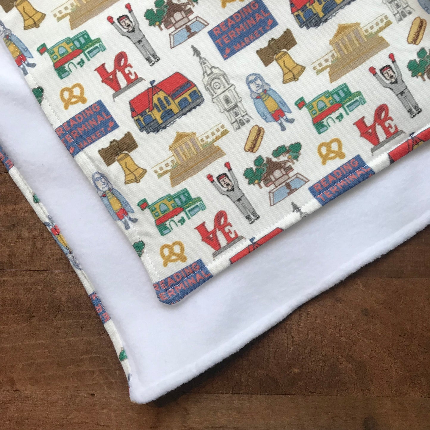 Fabric with a colorful pattern of iconic Philadelphia symbols and landmarks depicted on an organic cotton Ana Thorne Philly Baby Blanket displayed on a wooden surface.