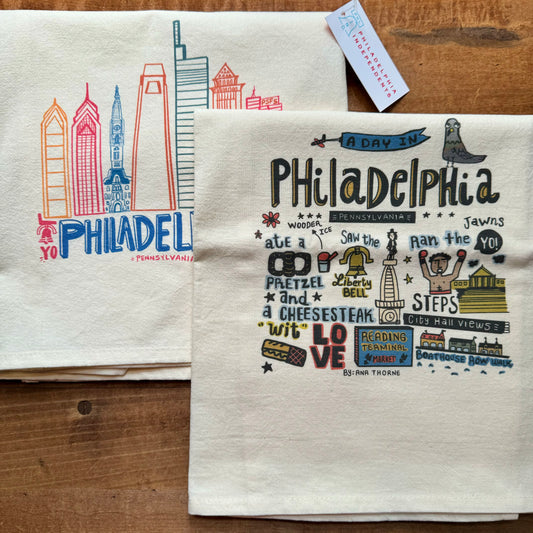 Two Philadelphia Tea Towels by Ana Thorne on a wooden surface make a great housewarming gift; one features colorful sketches of Philadelphia landmarks, and the other lists popular Philly attractions and slang.