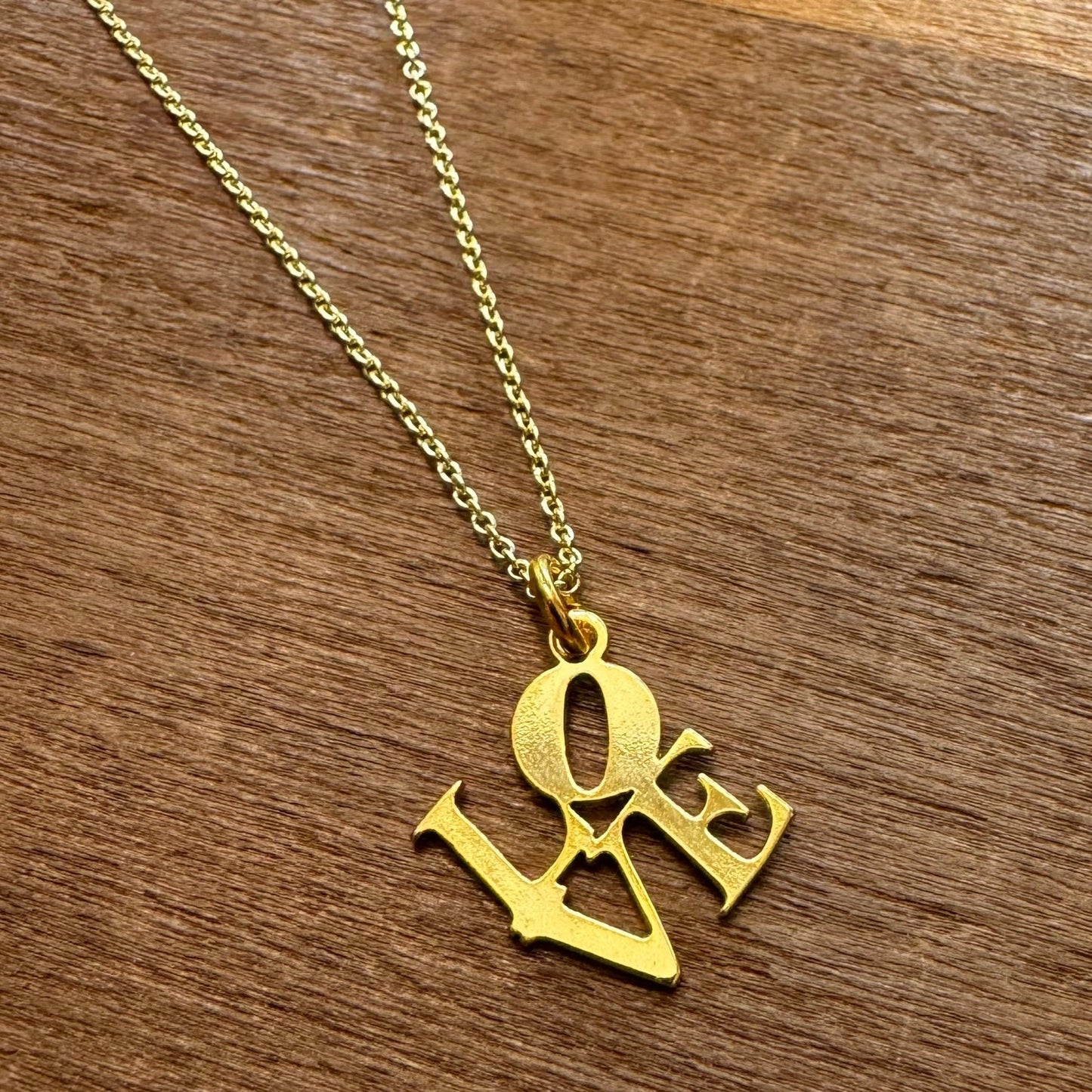 The LOVE Necklace by GeoMetricGem, a gold-plated piece featuring a pendant that spells out "LOVE" in uppercase letters, elegantly displayed on a wooden surface, epitomizes sophistication and elegance.