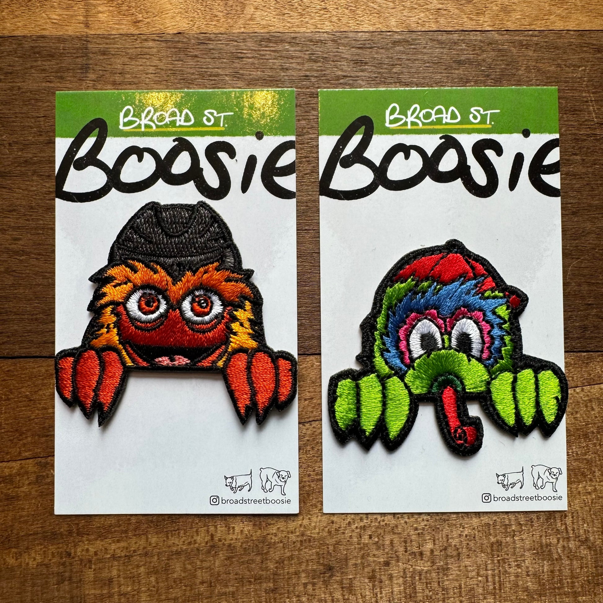 Two Gritty & Phanatic Patches on a wooden surface; the left patch depicts an orange monster with big eyes, and the right patch shows a multicolored monster, both featuring "Broad Street Boosie" text.