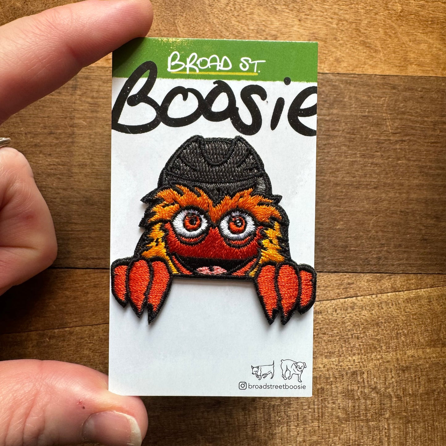 An embroidered patch featuring "Gritty & Phanatic Patches," an orange monster with big red eyes and a black hat, displayed on a wooden surface.