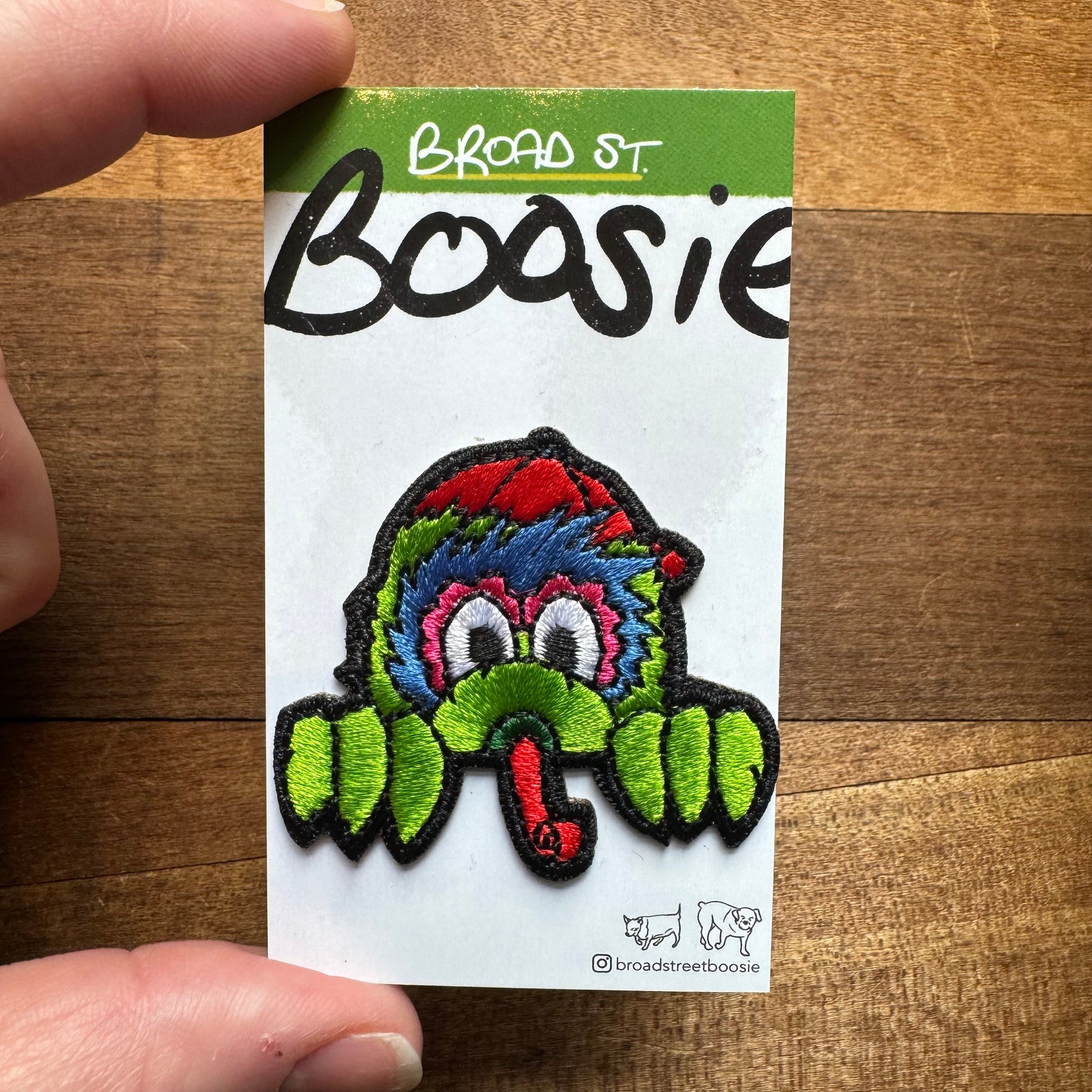 A hand holds a card with "Gritty & Phanatic Patches" written on it, featuring a colorful embroidered patch of a whimsical, green creature with a red tongue from Broad Street Boosie.