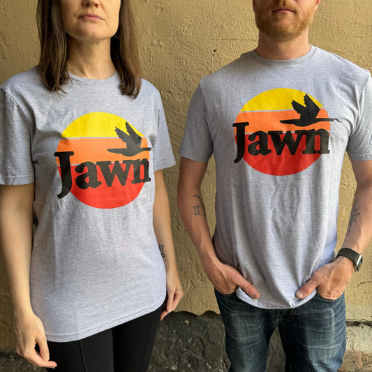 Two people stand against a beige wall wearing grey t-shirts from South Fellini, featuring a sunset graphic and the Philly slang word 