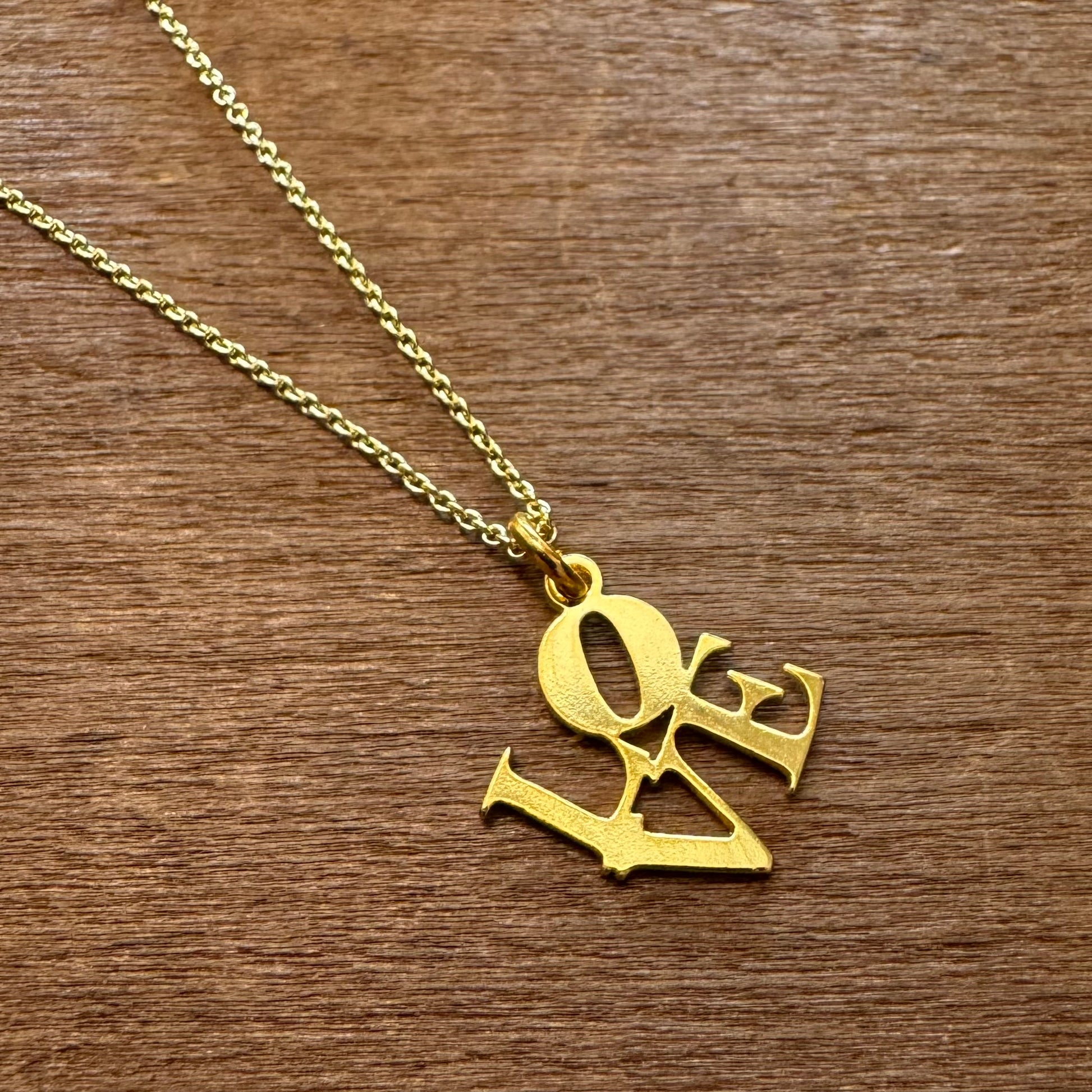 GeoMetricGem's gold-plated LOVE Necklace, featuring a pendant that spells out "love" in a stylized font, is elegantly showcased on a wooden surface.