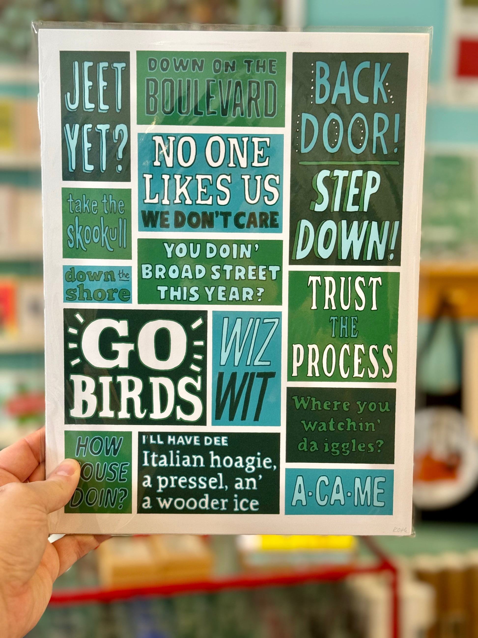 A person holds a sign with various Philly Lingo phrases in different fonts and colors, including "NO ONE LIKES US," "GO BIRDS," and "WIT WIT." The background includes shelves adorned with Kate Otte's Philadelphia Prints.
