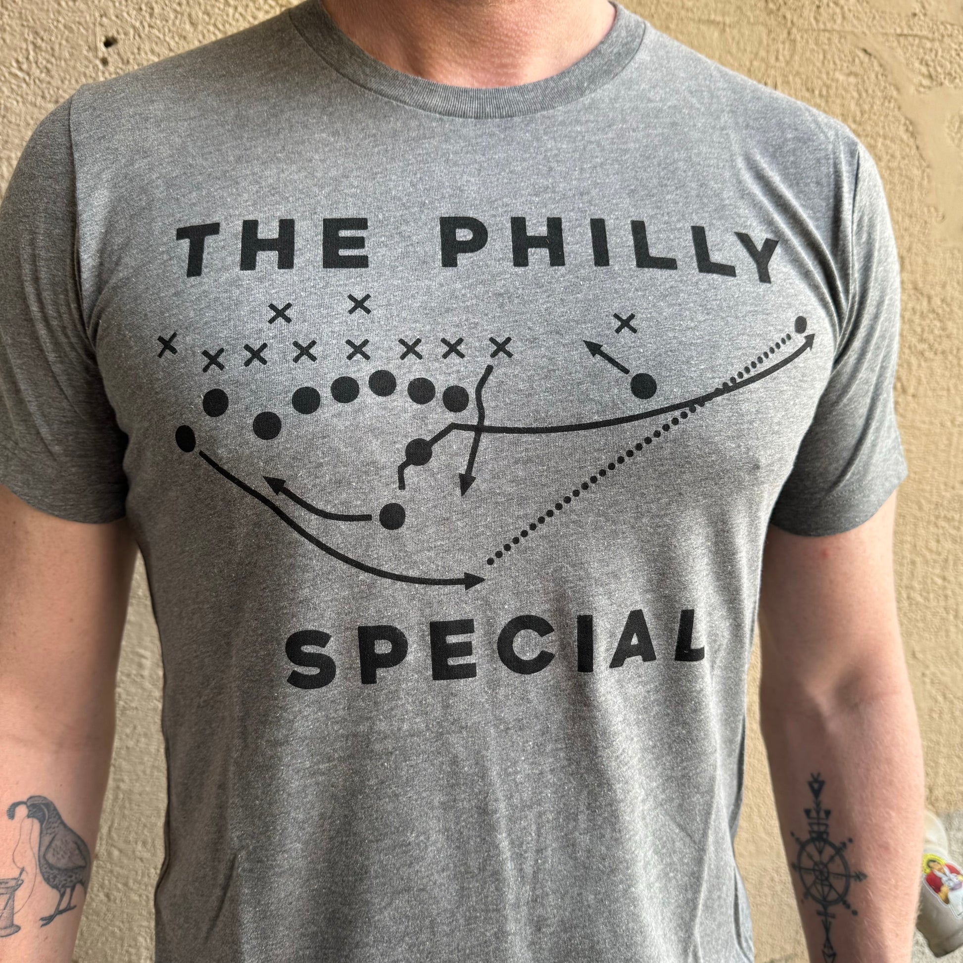 A person wearing a Philly Special T-Shirt by exit343design, featuring a football play diagram labeled "The Philly Special." The grey shirt is Super Bowl-inspired with markings including Xs, Os, and arrows. The person's torso and arms are visible.