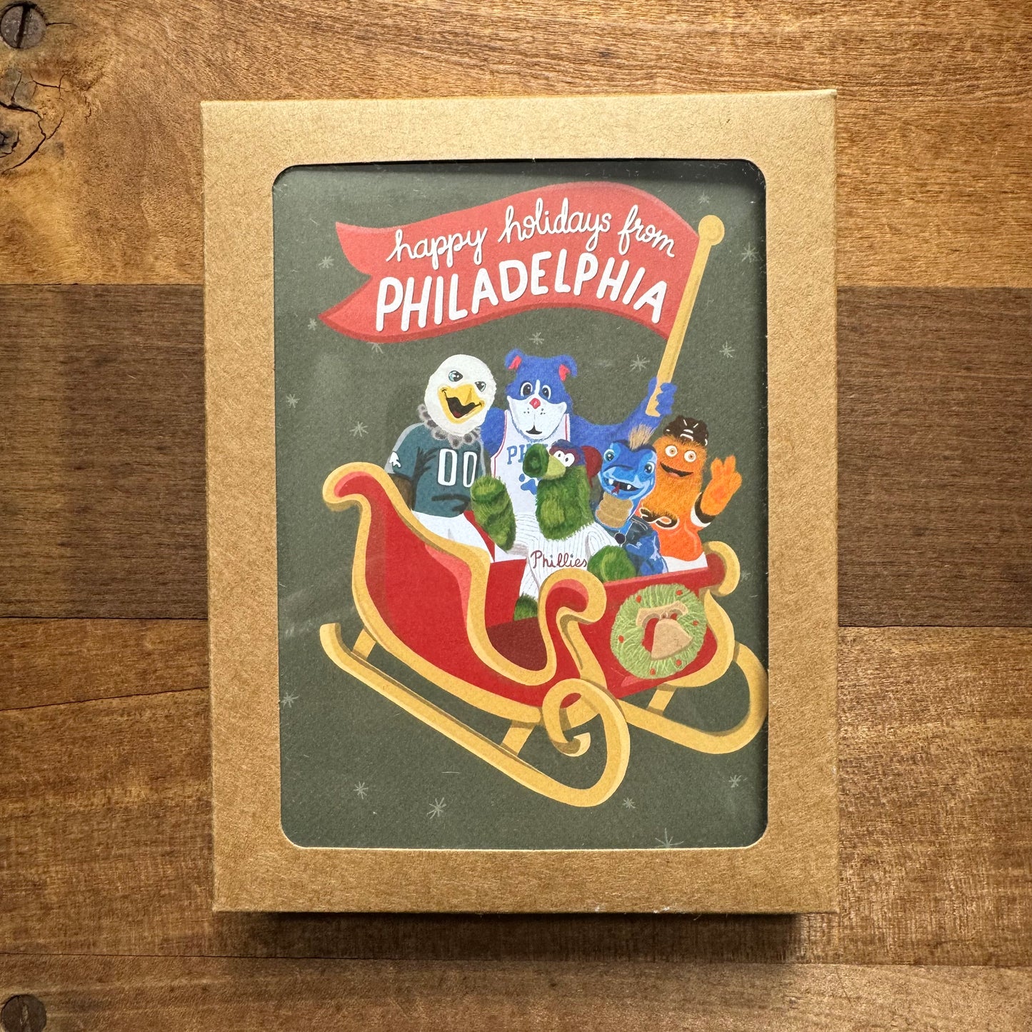 The Holiday Card Packs II by Parcel Island feature a holiday card with cartoon mascots in a sleigh and the text "Happy Holidays from Philadelphia" on a wooden background, making it perfect for pairing with envelopes to send your festive greetings.