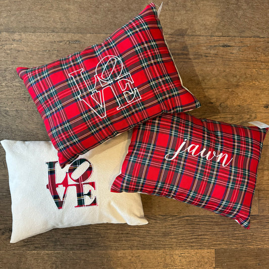 Three festive "Holiday Plaid Pillows" by The Pillow Works rest on a wooden surface; two are red plaid with "LOVE" and "Jawn" embroidered, and one is white with a plaid LOVE design.