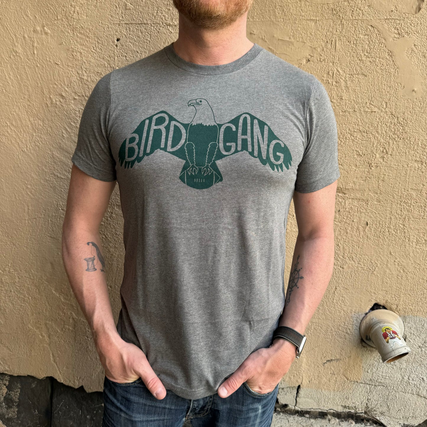 A person wearing a gray unisex exit343design Bird Gang T-Shirt with a graphic of an eagle and the text "Philly’s Bird Gang" stands against a textured beige wall. Their hands are resting in their jeans pockets.