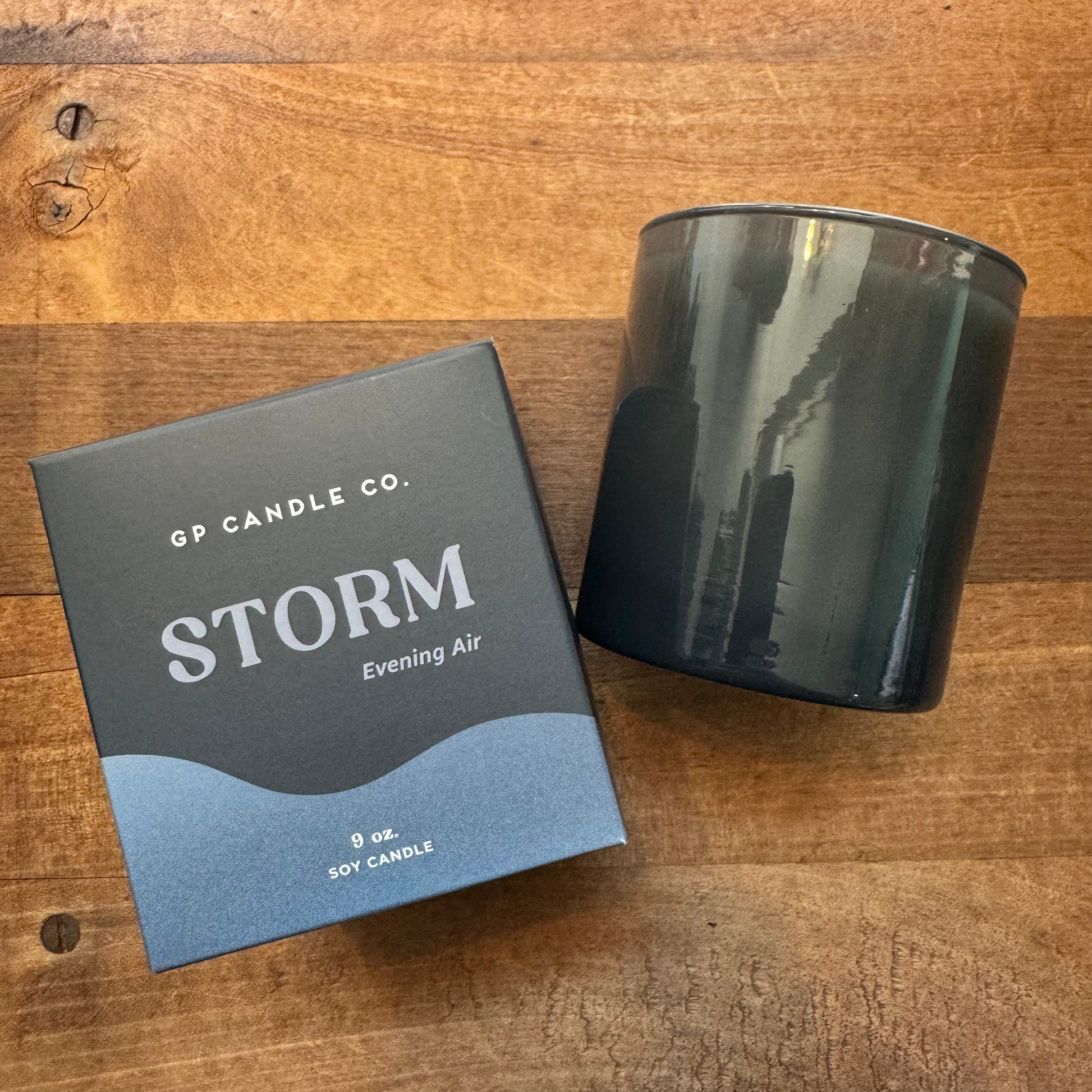 A black candle jar with its lid beside a box labeled "GP Candle Co. Soy Candles-Hue Collection, Storm Evening Air, 9 oz," is displayed on a wooden surface, showcasing the fresh scents of hand-poured soy wax candles.