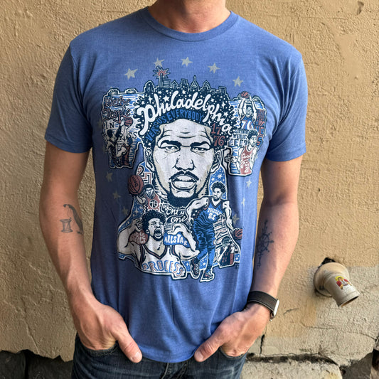 A person is wearing a blue Sixers Joel Embiid T-Shirt, featuring an illustrated design with the text "Philadelphia" and various basketball-themed graphics. The unisex Paul Carpenter shirt showcases only the torso and arms of the person.