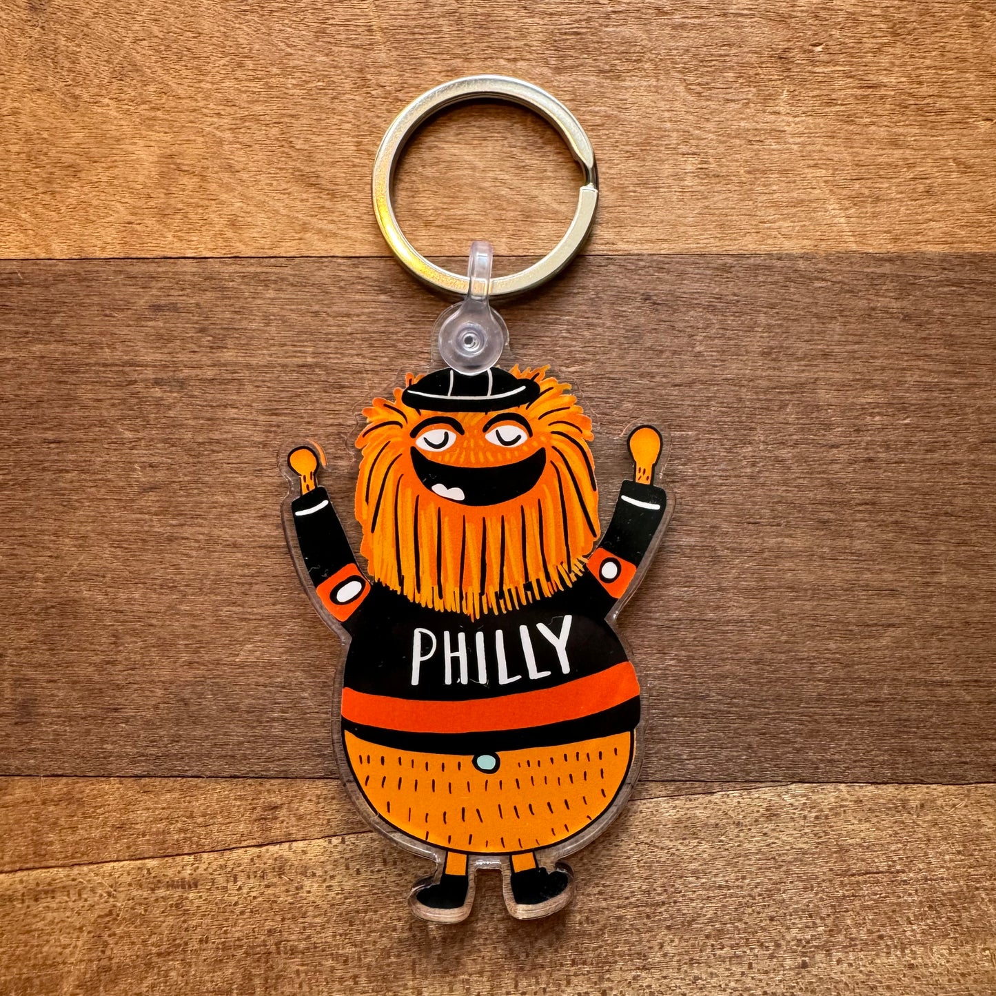 Keychain featuring an orange, fluffy character with raised arms, wearing a black shirt that says "PHILLY" and a beanie. This Gritty & Phanatic keychain by Ana Thorne is placed on a wooden surface, capturing the essence of Philly spirit.