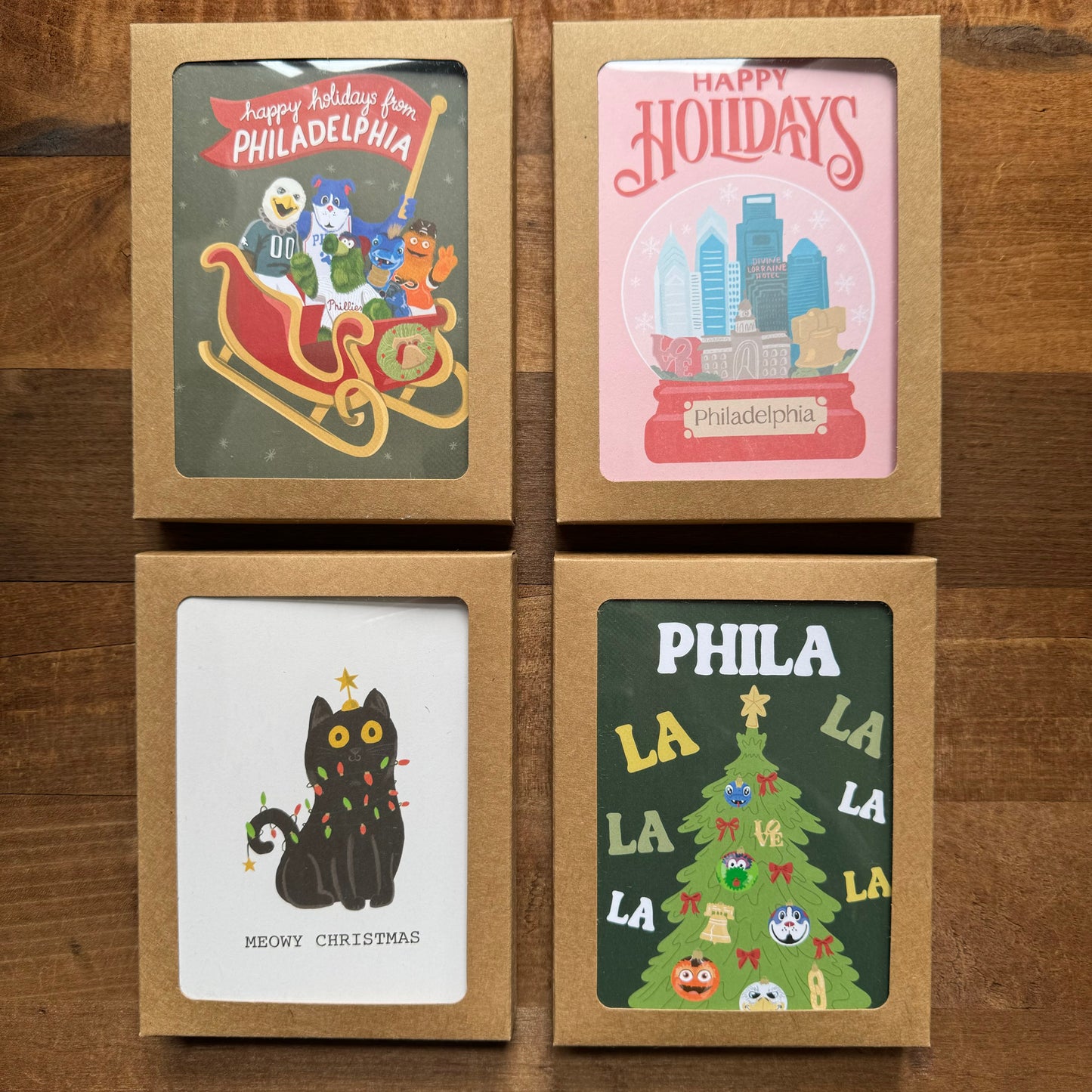 Introducing the Holiday Card Packs II by Parcel Island: Four holiday-themed Philly cards beautifully presented in brown boxes. These festive cards feature designs celebrating Philadelphia, with illustrations of a sleigh, city skyline, black cat, and a decorated Christmas tree. Each blank card comes complete with an envelope for your convenience.