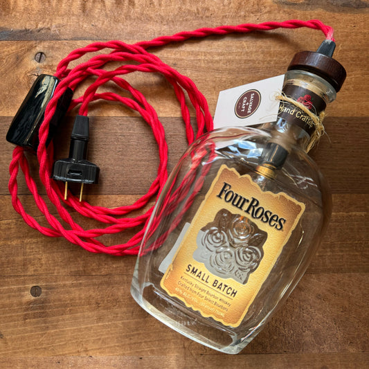 A Salvage & Craft Bottle Pendant Lamps repurposed into a whiskey lamp, featuring a red twisted fabric cord and a black plug, resting on a wooden surface and crowned with an Edison bulb.