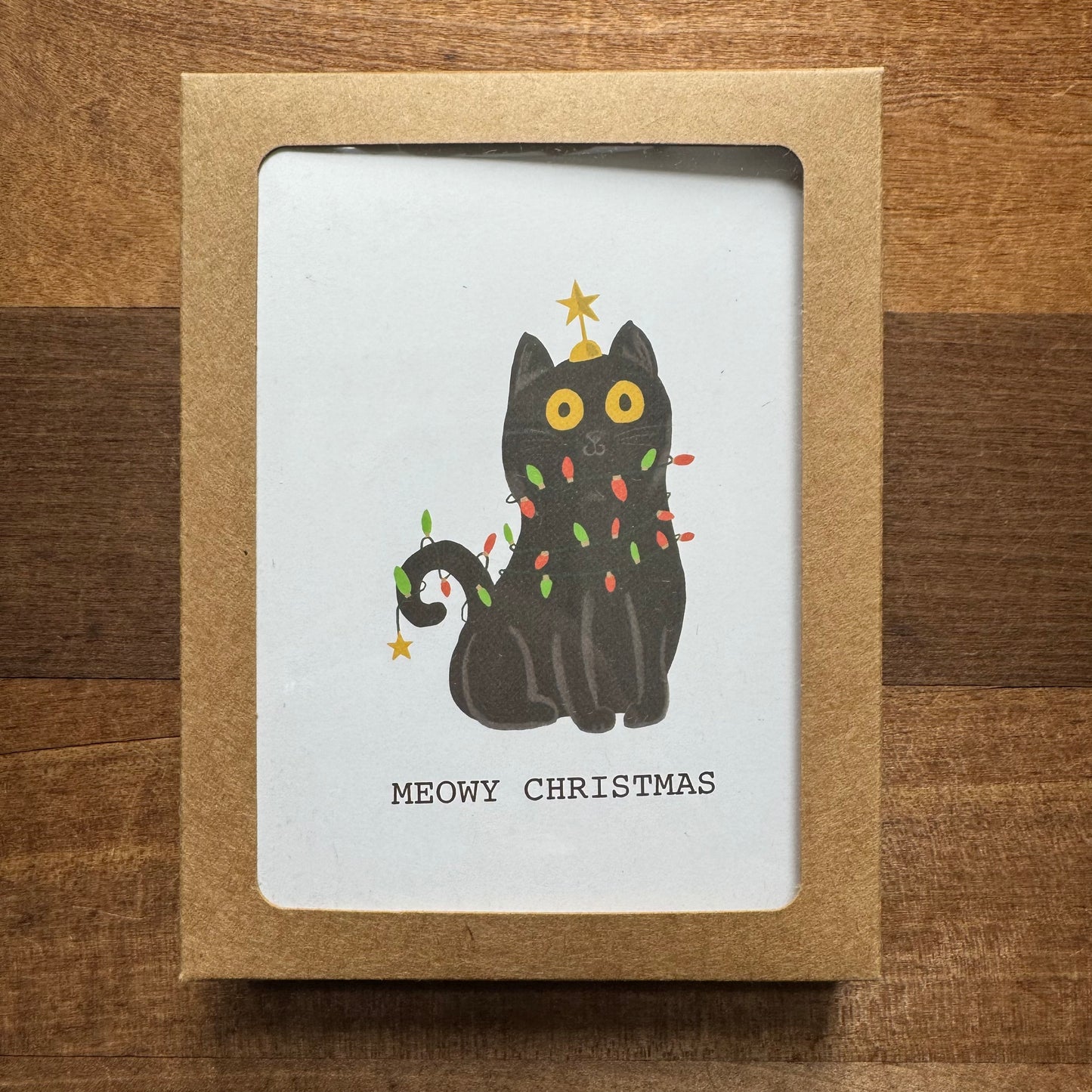 The Holiday Card Packs II by Parcel Island feature a charming black cat illustration entwined in Christmas lights, with a star on its tail and the text "MEOWY CHRISTMAS." These blank cards allow for personalized messages and come with matching envelopes, all beautifully displayed on a wooden surface.