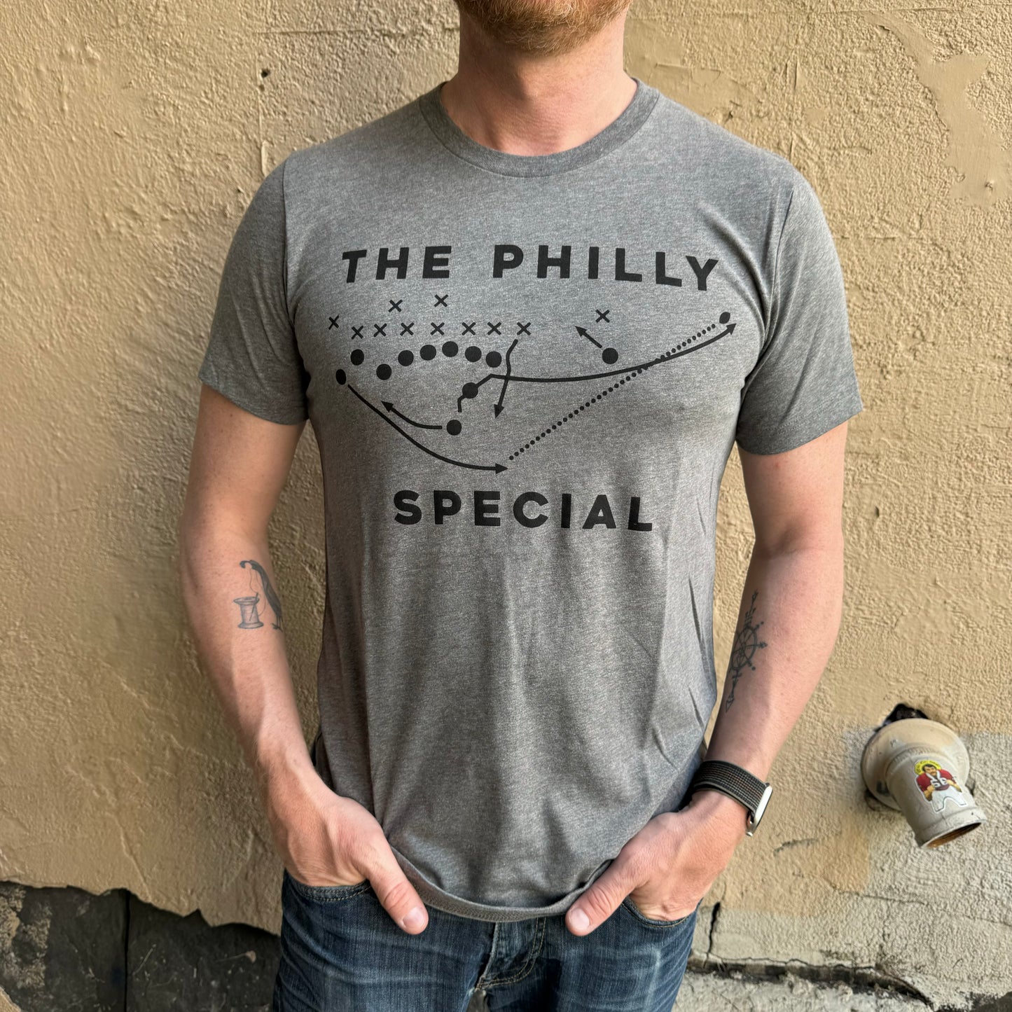 A person wearing a Philly Special T-Shirt, inspired by the Super Bowl, stands against a textured beige wall. The shirt, from the brand exit343design, features a football play diagram with the text "The Philly Special." The person has tattoos on their forearms and their hands in their pockets.