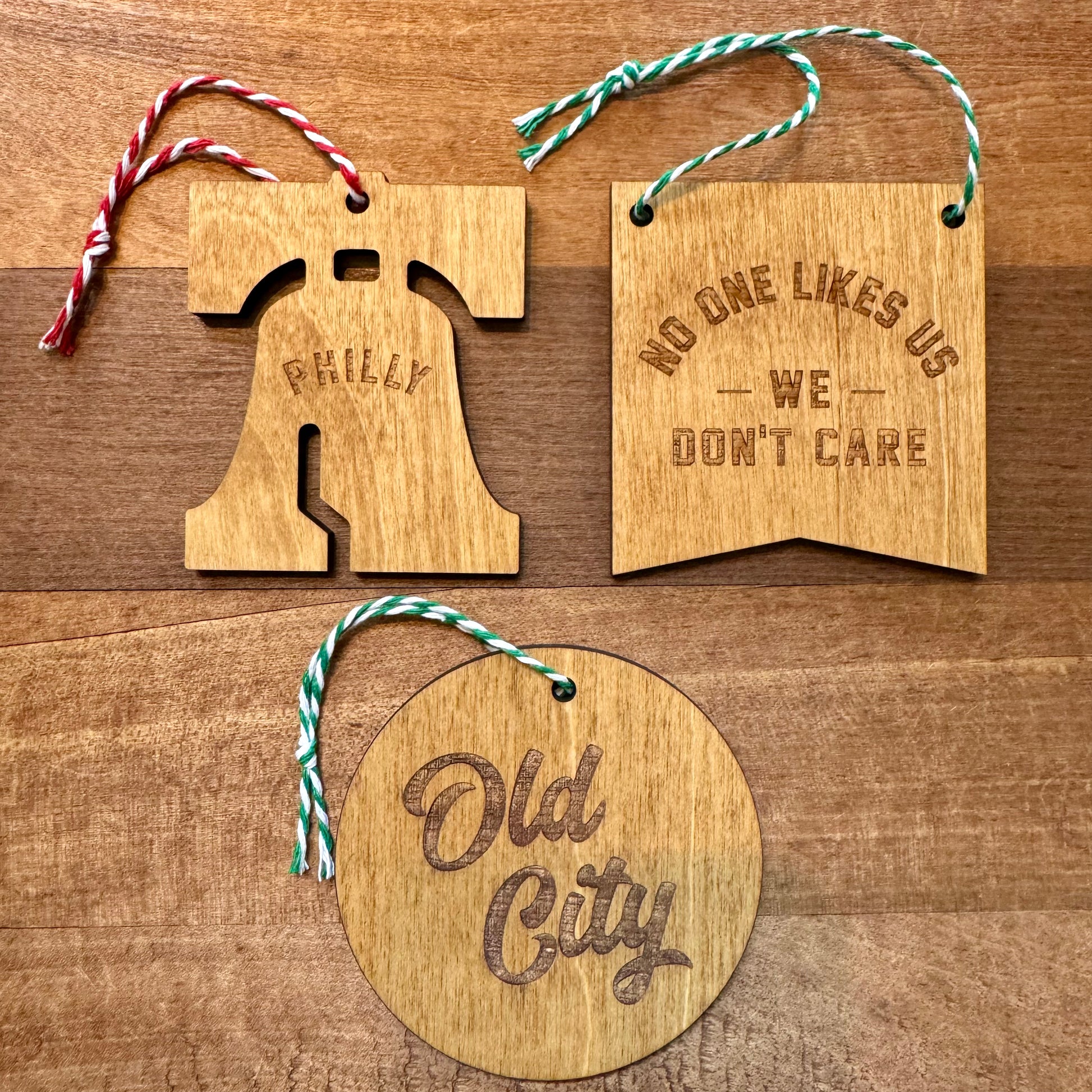 Frankadelphia's Philly Wood Ornaments feature three Philly-themed designs: one laser-etched wood shaped like the Liberty Bell with "Philly," one rectangular with "No One Likes Us We Don't Care," and one round with "Old City," all adorned with colored strings.