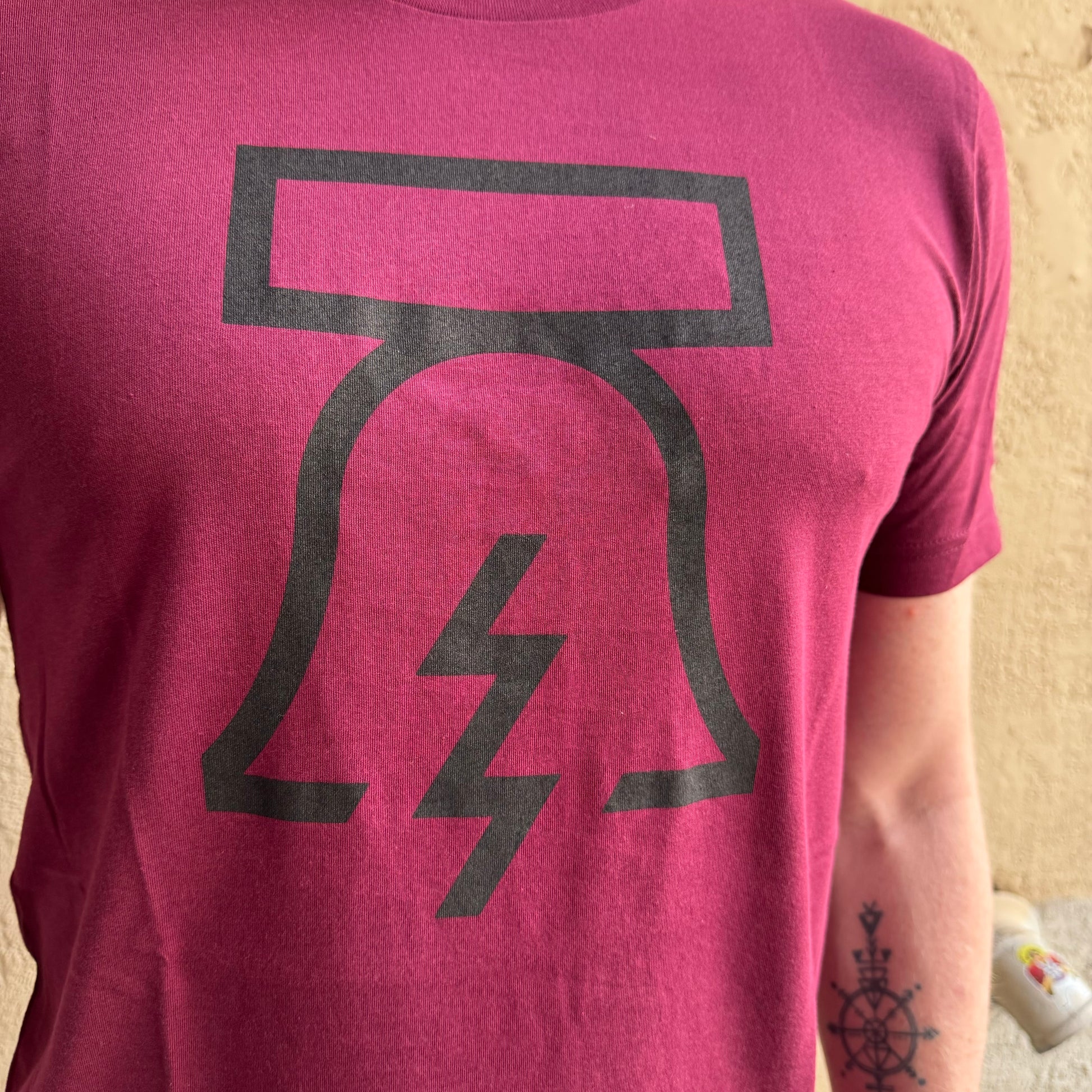 Close-up of a person wearing a maroon unisex Philadelphia Independents Bell & Bolt T-Shirt with a black, stylized design of the Liberty Bell and lightning bolt. The person has a tattoo on their left forearm.