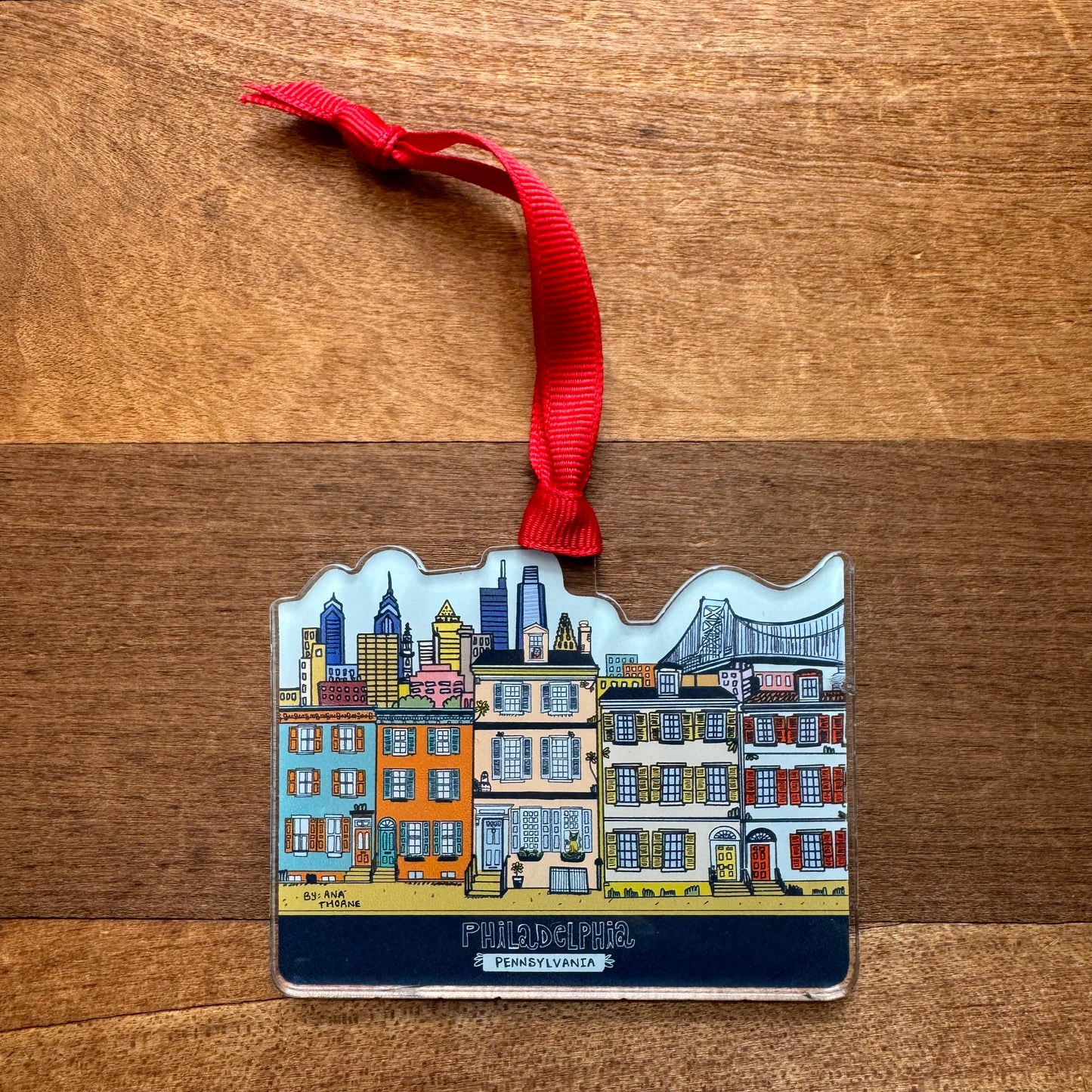 The Philly Acrylic Ornaments by Ana Thorne embody true Philly pride, showcasing a Philly Rowhome, city buildings, and a bridge. Each ornament comes with a red ribbon for hanging.