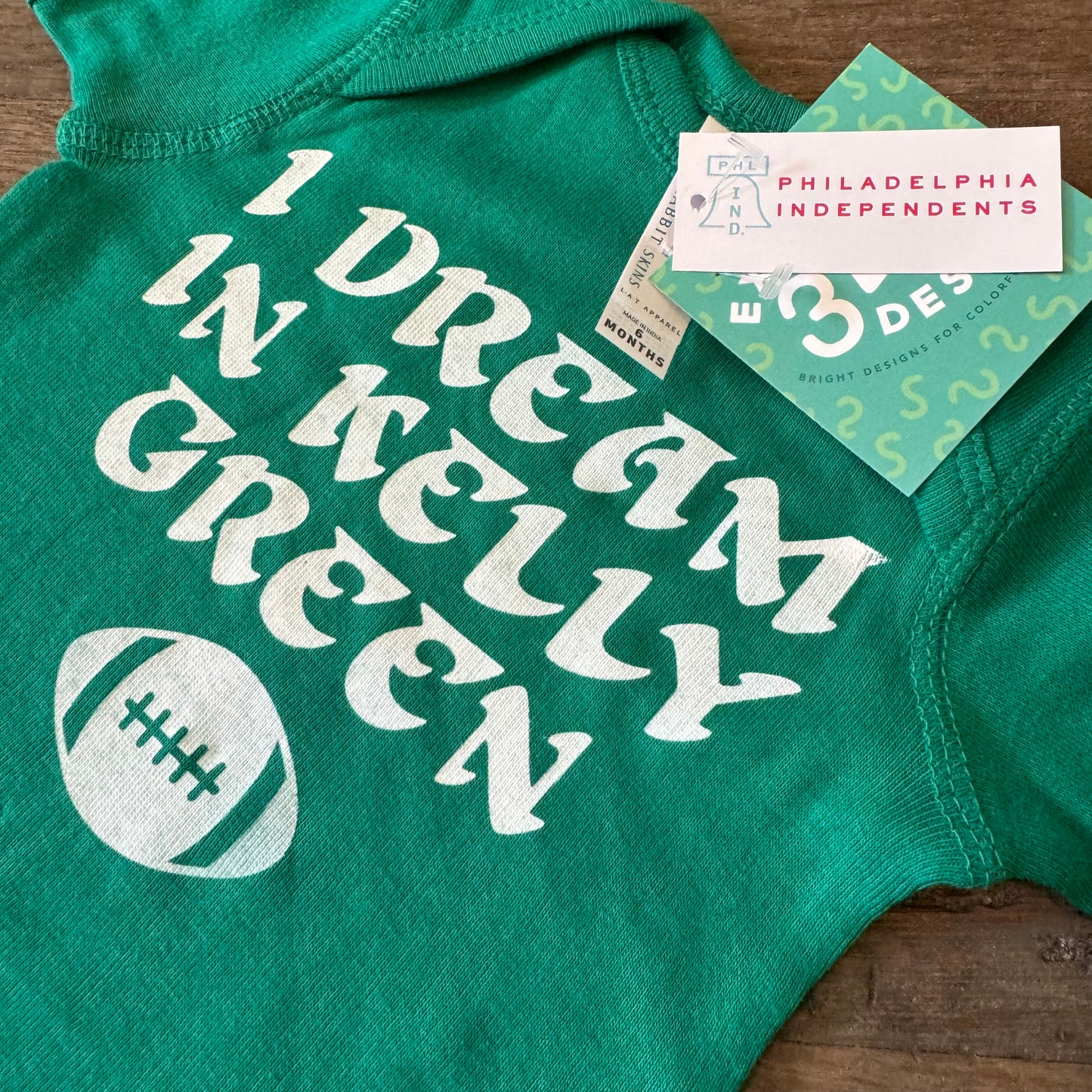 The "I Dream in Kelly Green Baby Onesie" by exit343design features a football graphic and the phrase "I Dream in Kelly Green." Made from 100% cotton, it comes with a product tag and a professionally printed card from Philadelphia Independents.