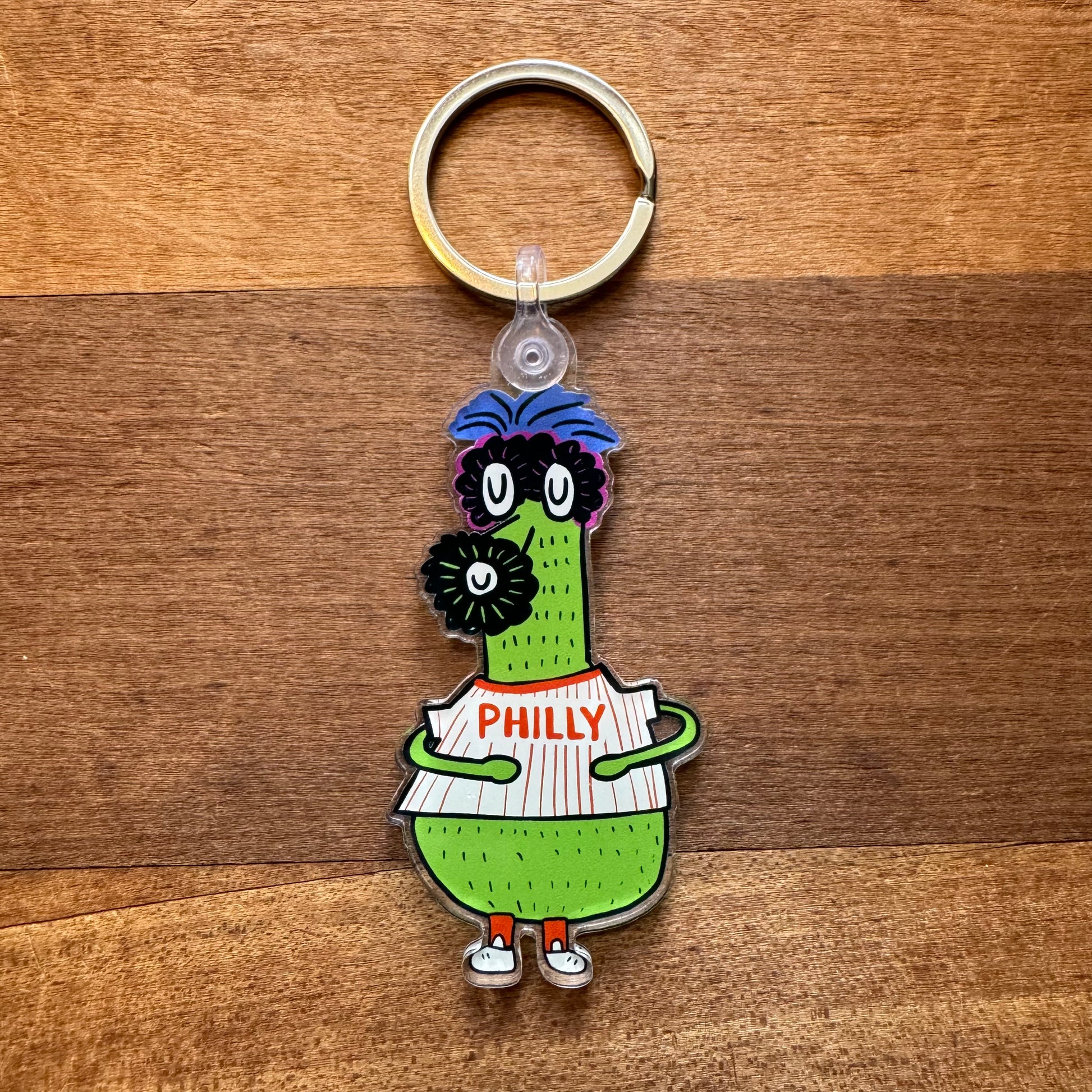 A keychain from Ana Thorne's Gritty & Phanatic collection, showcasing a green cartoon character holding a sign with "PHILLY," stands on a wooden surface.