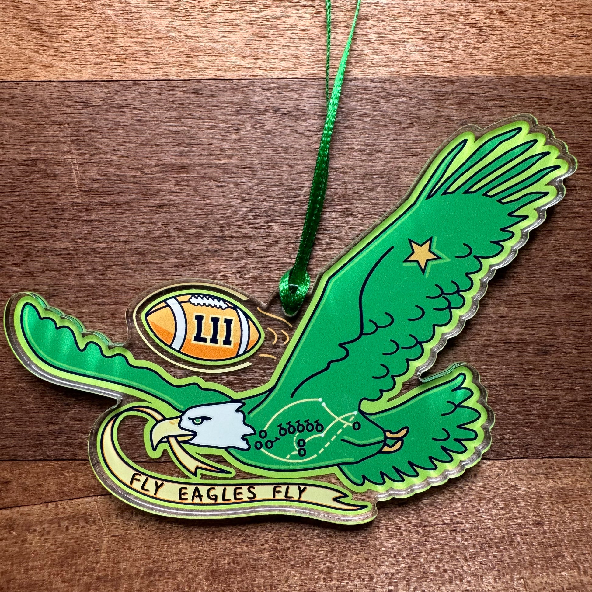 The Philly Acrylic Ornaments III by Jeffrey Arden Art feature a Philly sports-themed design with an illustrated green eagle holding a football marked "LII." It includes a green ribbon for hanging and has the text "Fly Eagles Fly" on a banner beneath the eagle, showcasing Jeffrey Arden Art's signature style.