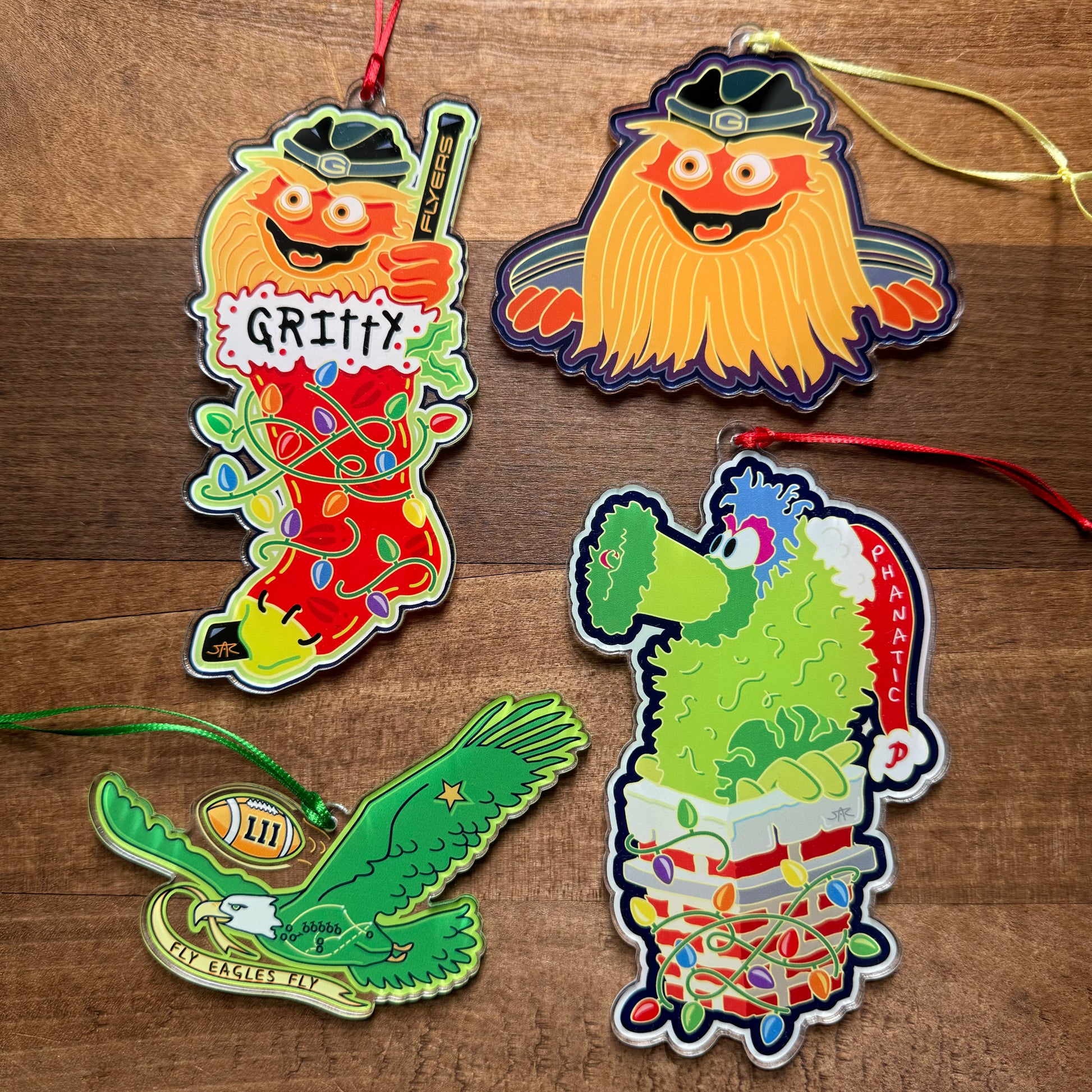 Four vibrant Philly Acrylic Ornaments III, featuring animated sports mascots and a decorated football eagle designed by Jeffrey Arden Art, are displayed on a wooden surface.