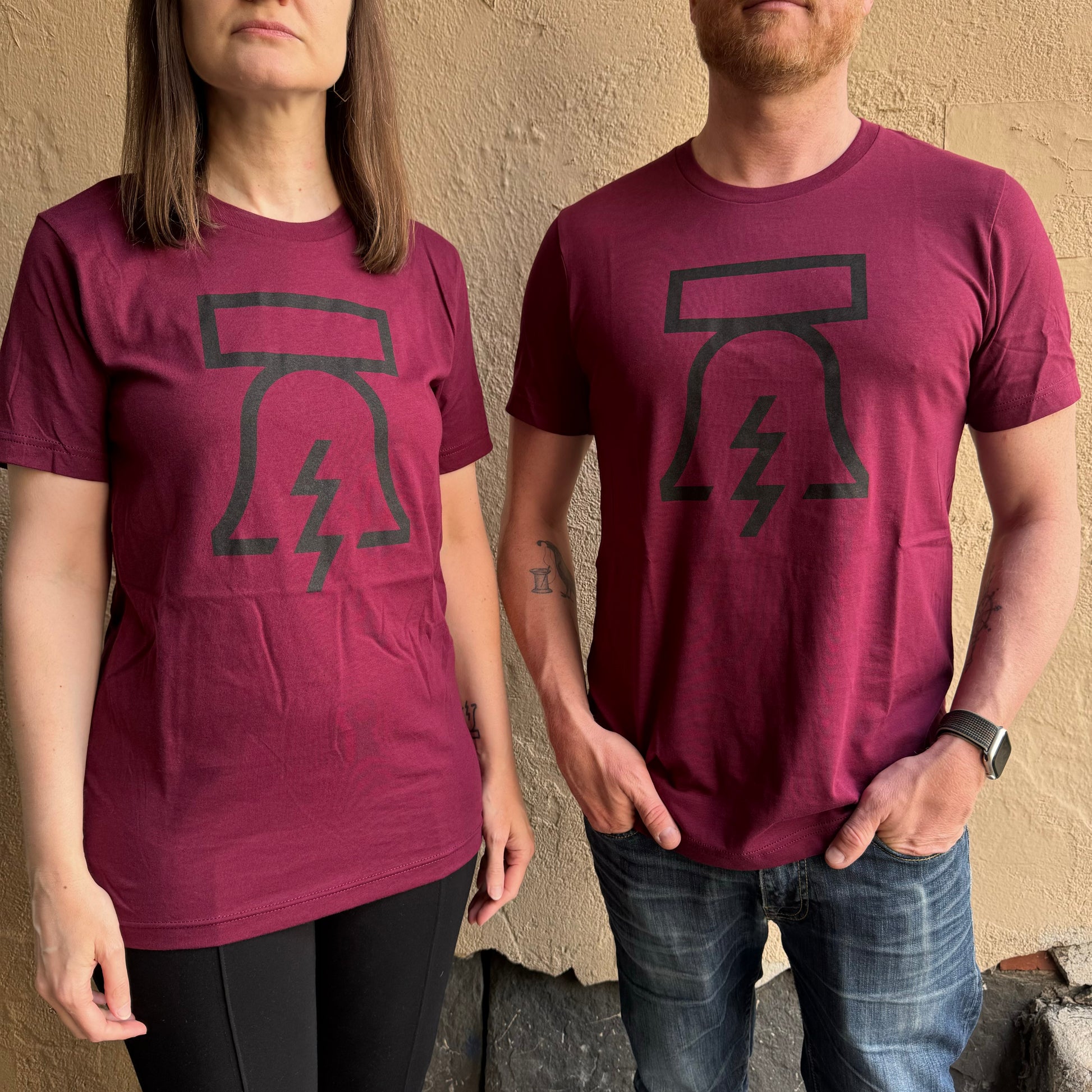 Two people stand side by side against a textured wall, both wearing matching unisex Philadelphia Independents maroon Bell & Bolt T-Shirts with a black graphic of a cracked Liberty Bell with a lightning bolt.