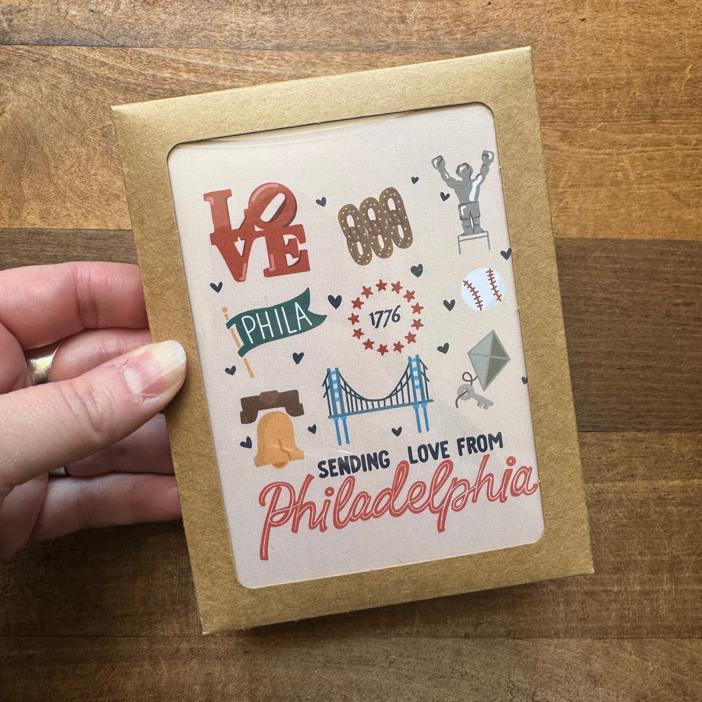 A hand holds a greeting card from the Parcel Island Philadelphia Notecard Pack, showcasing various Philadelphia-themed illustrations like the LOVE sculpture, Liberty Bell, Independence Hall, sports symbols, and the phrase "Sending Love from Philadelphia." This charming card is part of a delightful pack that truly captures Philly phlair.