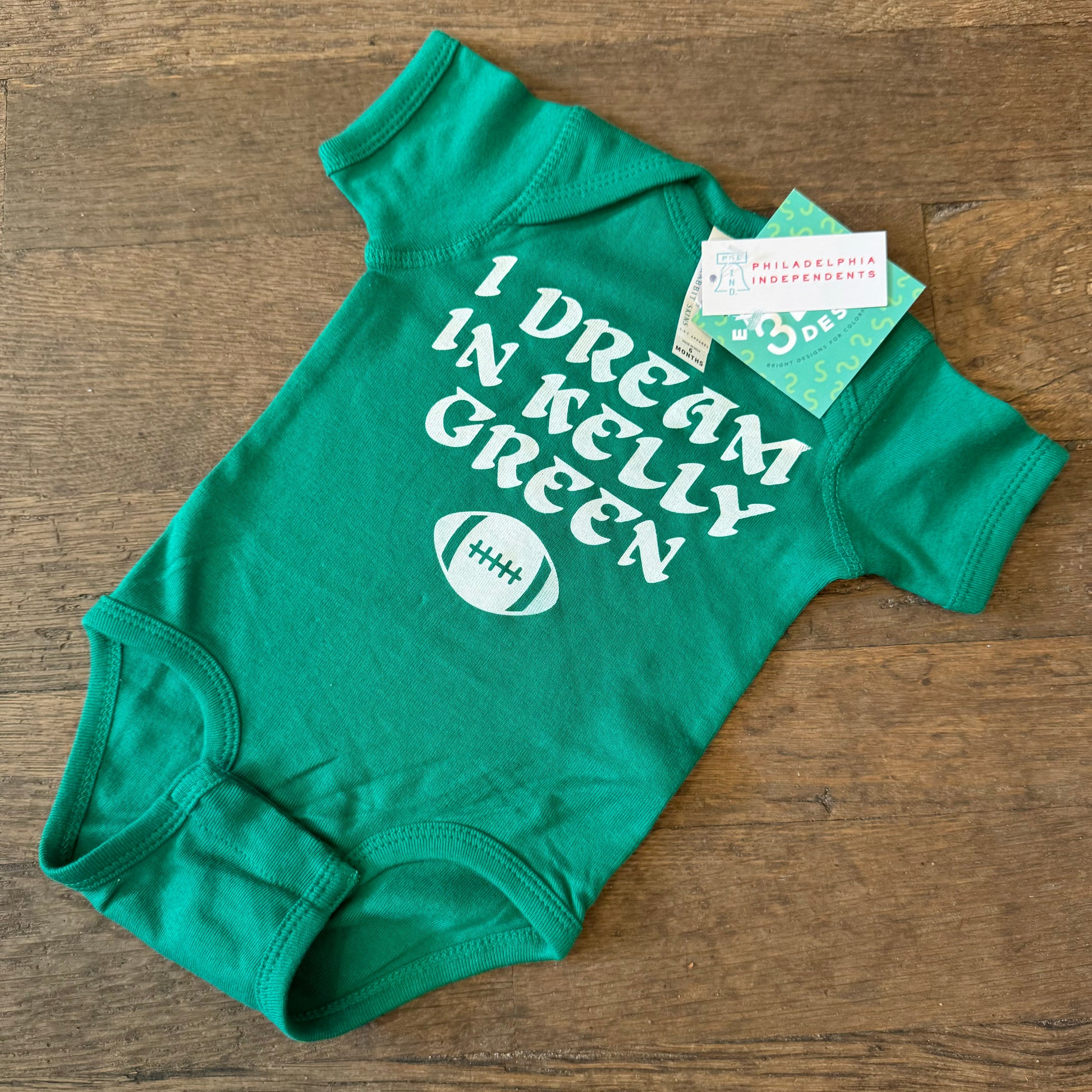 The I Dream in Kelly Green Baby Onesie by exit343design, made entirely of cotton, features the text "I DREAM IN KELLY GREEN" along with a small football graphic. This Kelly Green onesie, complete with tags attached, is laid out on a wooden surface.