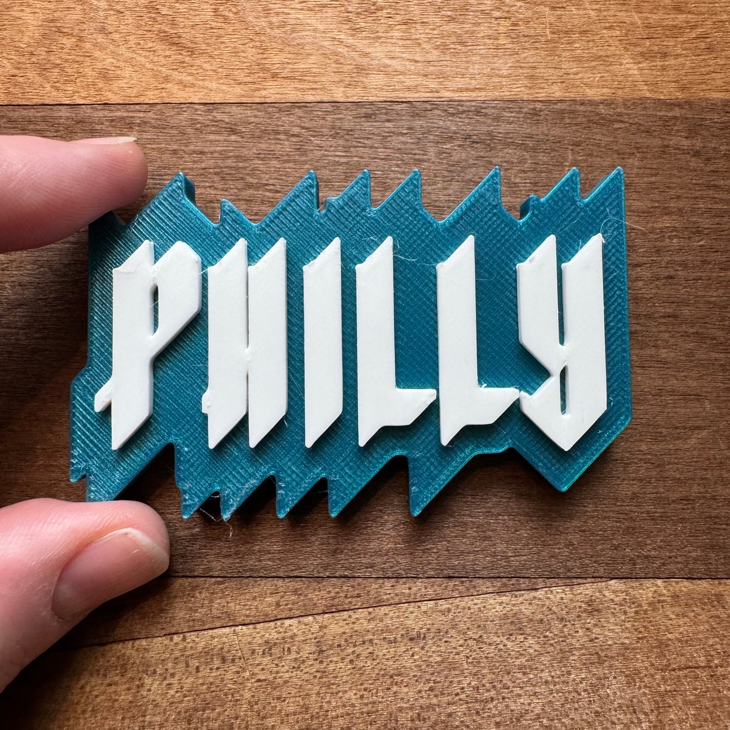 A hand holding a blue and white 3D-printed object with the word "PHILLY" on it, placed on a wooden surface. This could be one of those Rosewood Home Philly Magnets that capture the city's essence in miniature.