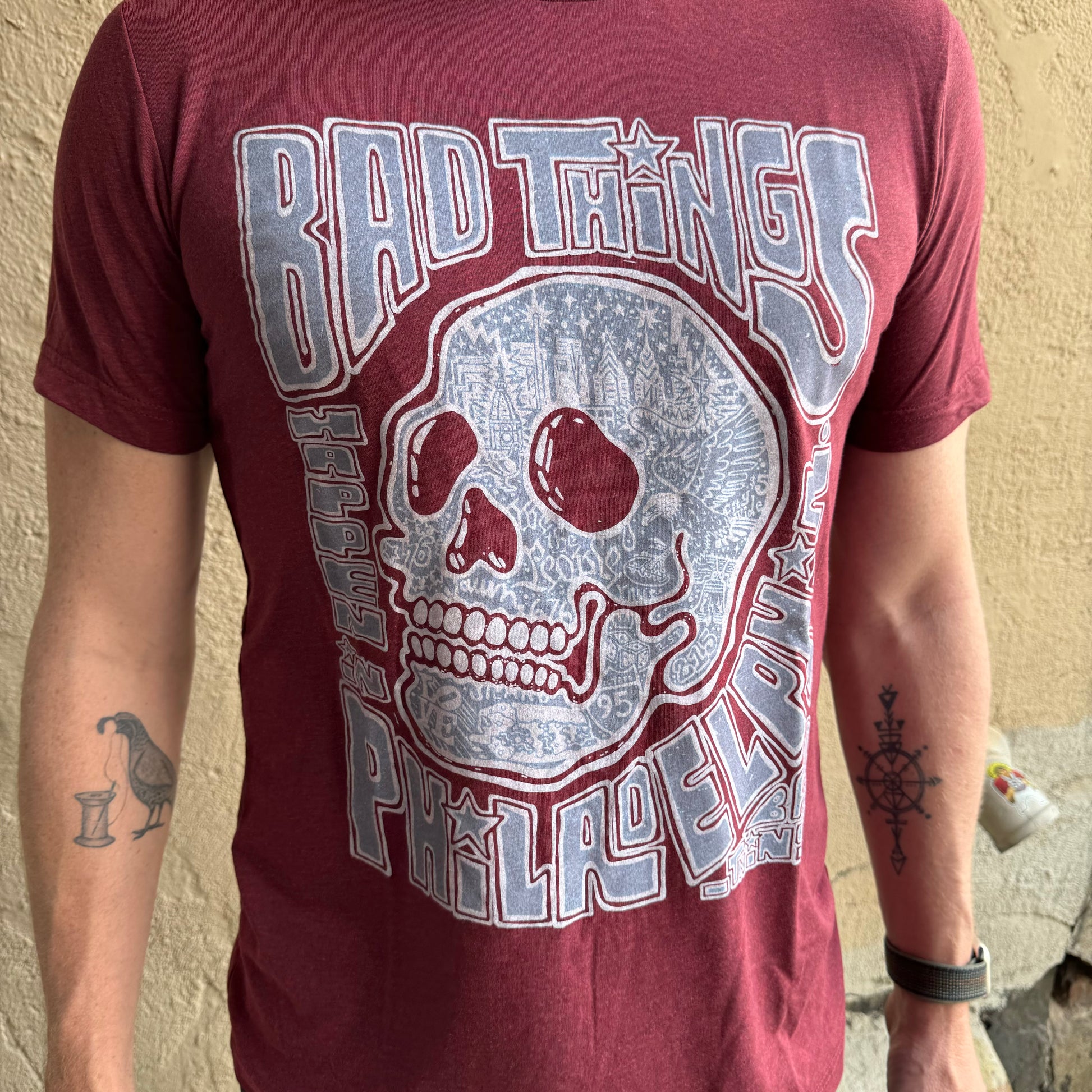 A person wearing a super soft Paul Carpenter maroon Bad Things Happen T-Shirt with a graphic design featuring a skull and text that reads "Bad Things Happen in Philadelphia." The Paul Carpenter shirt showcases the person's arms adorned with multiple tattoos.