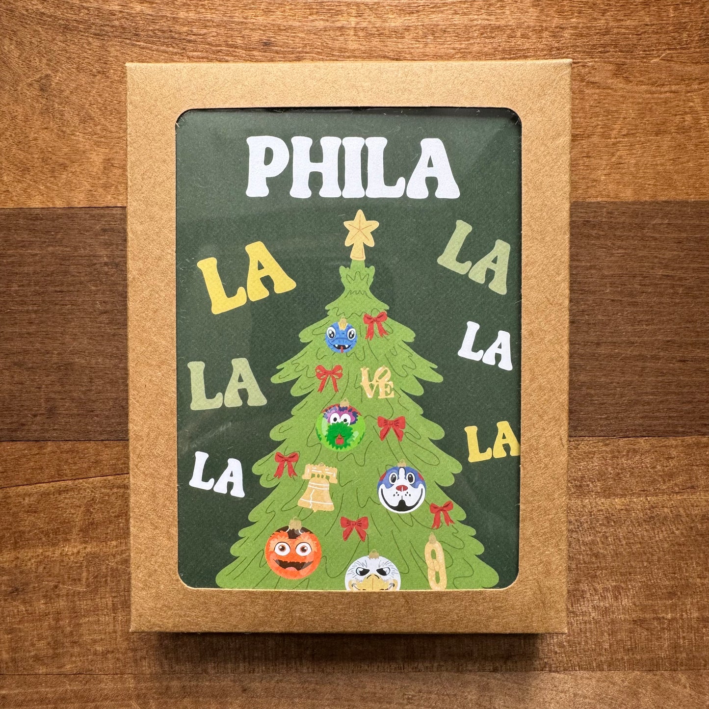 This Holiday Card Packs II by Parcel Island features a card adorned with a festive Christmas tree, decorated with an assortment of ornaments and inscribed with the playful text "PHILA LA LA LA LA." Ideal for fans of Philadelphia-themed greeting cards, the card is set against a charming wooden background and comes complete with matching envelopes for an all-inclusive holiday greeting experience.