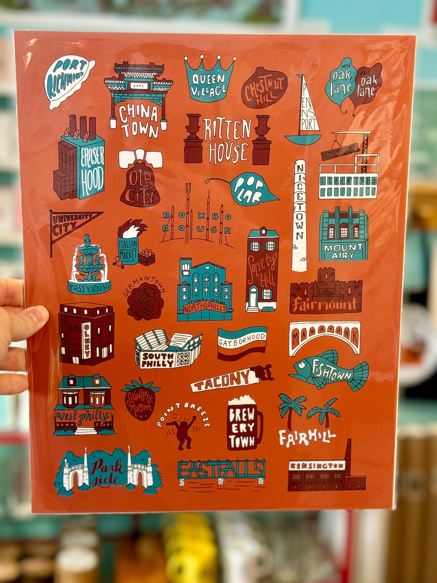 A hand holds a vibrant Philadelphia Prints by Kate Otte, showcasing the illustrations and names of various neighborhoods and landmarks such as Rittenhouse, Chinatown, and Fishtown, all set against an orange background.