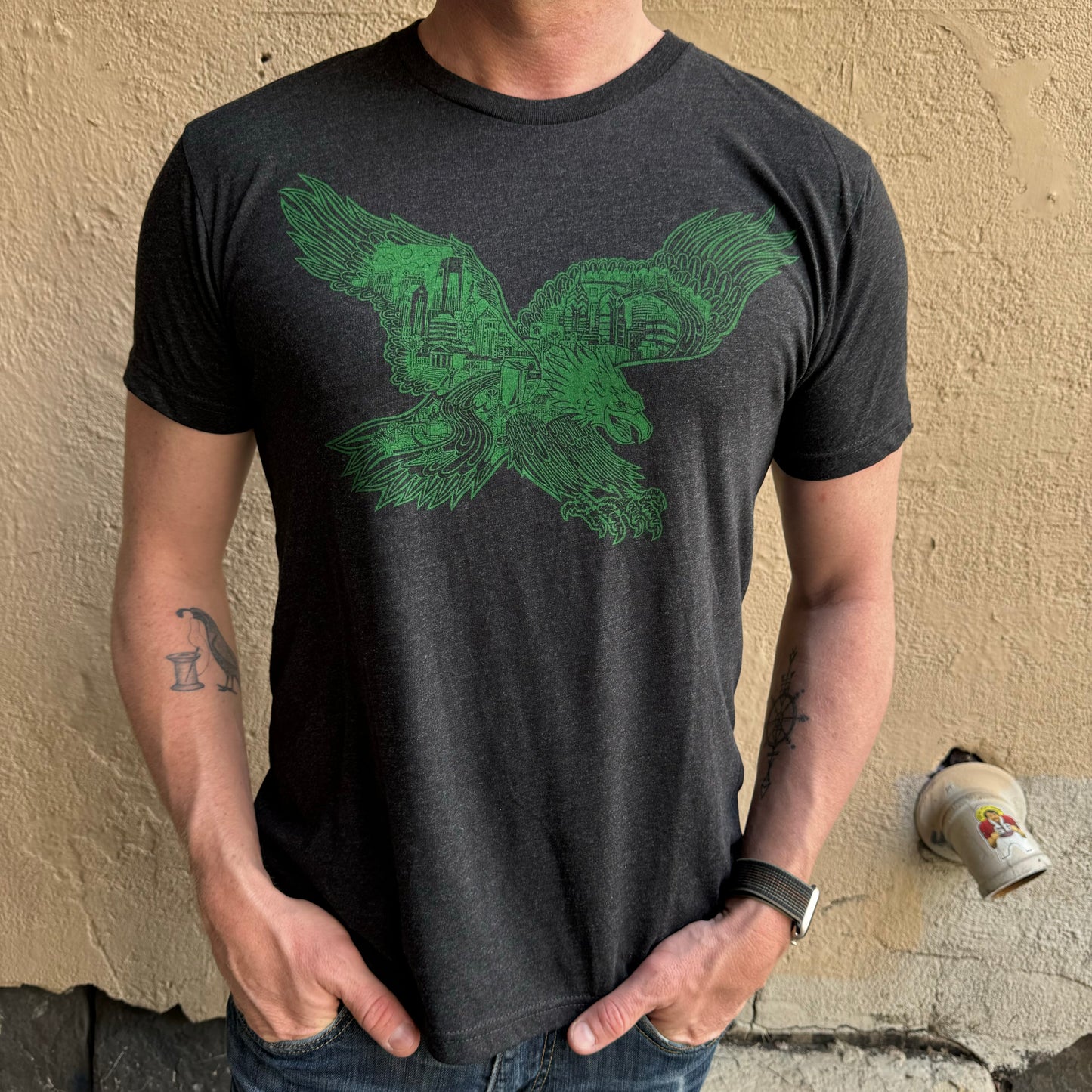 A person wearing a dark gray Swooping Eagle T-Shirt by Paul Carpenter stands against a beige textured wall with their hands in their pockets. They have tattoos on their forearms and are wearing a smartwatch, subtly blending urban fashion with personal style.