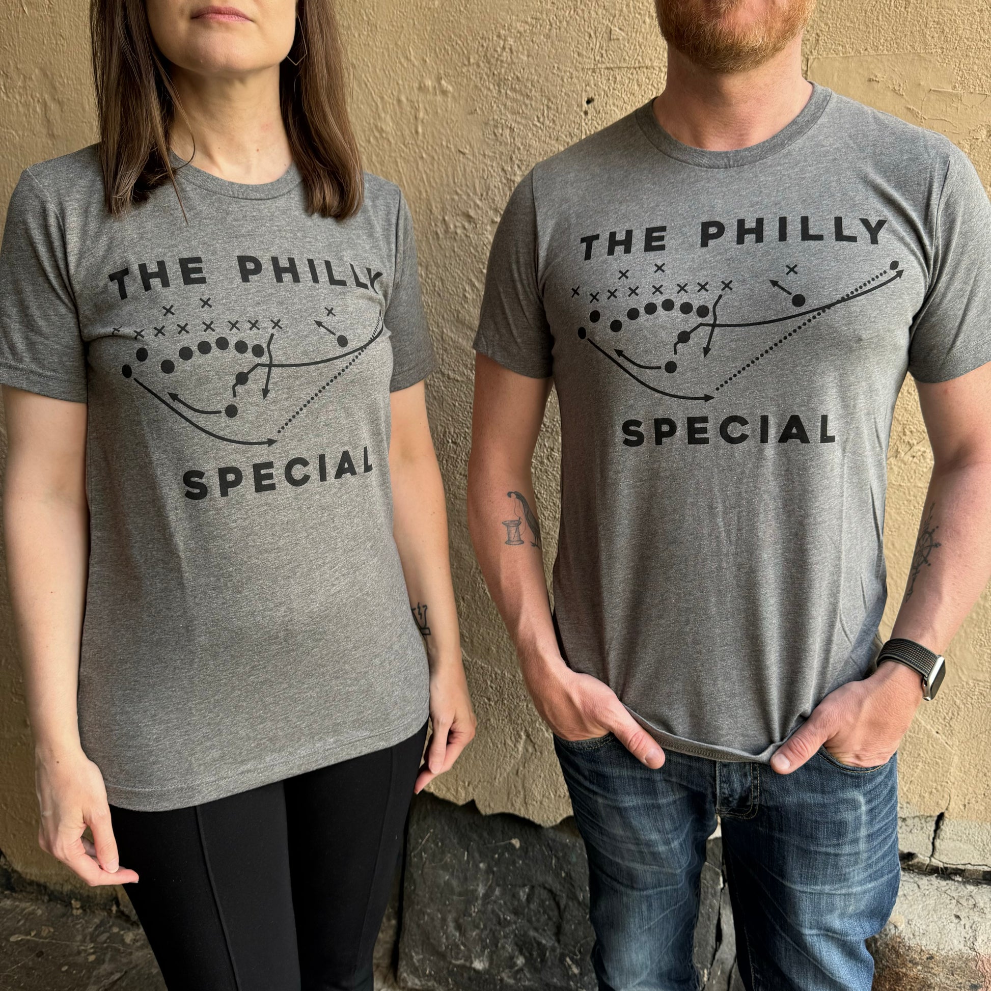 Two people, a woman and a man, wearing matching Philly Special T-Shirts in grey that feature a football play illustration, stand against a beige wall. These Super Bowl-inspired shirts are designed by exit343design.