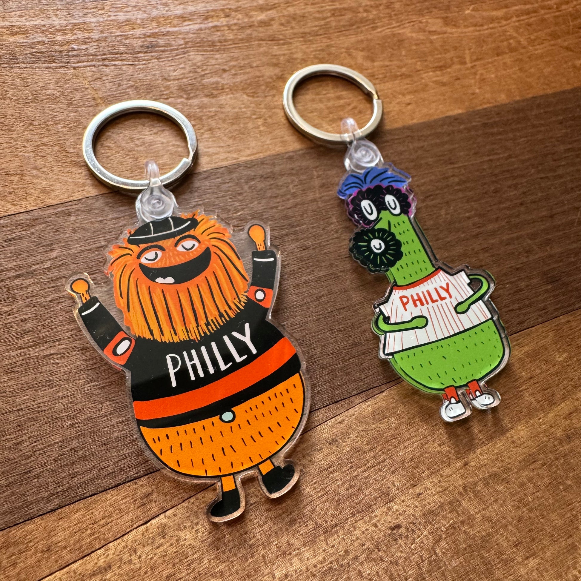 Two *Gritty & Phanatic Keychains* by *Ana Thorne* are on a wooden surface. One is an orange and black Gritty keychain sporting "PHILLY" on its shirt; the other is a green Phanatic keychain, wearing glasses and holding a "PHILLY" sign.