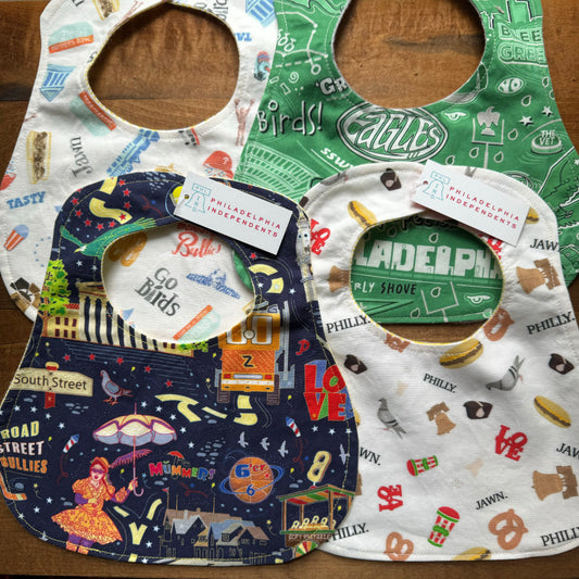 Philly Print Baby Bibs from Sew Much Cooler, featuring colorful Philadelphia-themed patterns, are displayed on a wooden surface. Each handmade bib is crafted from premium cotton print and comes with a tag from "Philadelphia Independents.