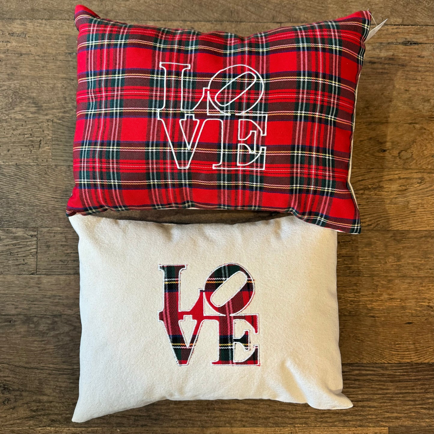 Two festive Holiday Plaid Pillows from The Pillow Works rest on a wooden floor; the top pillow features a red plaid pattern with "LOVE" embroidered in white, while the bottom pillow is beige with "LOVE" rendered in a combination of plaid and black lettering.