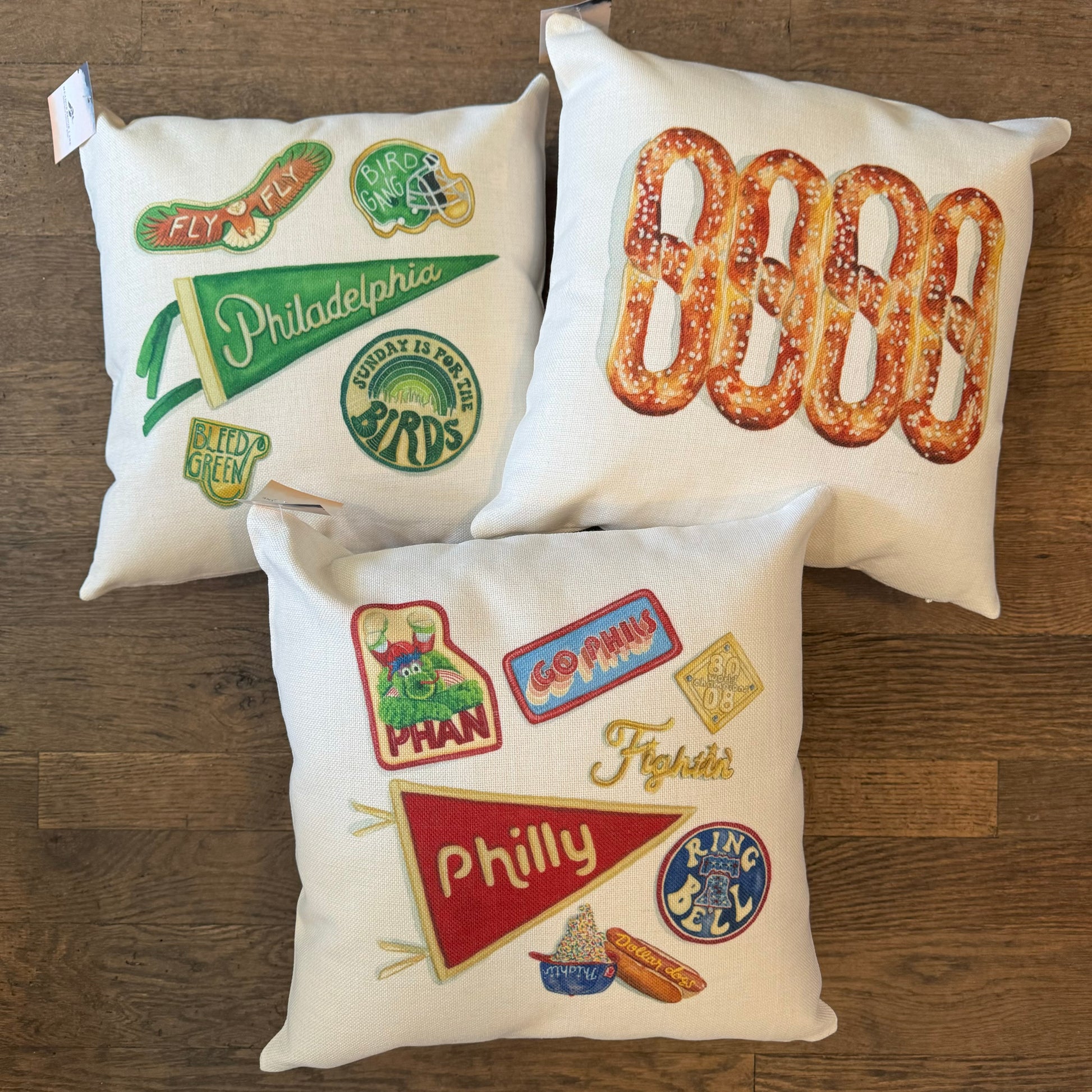 Three Philly Illustrated Pillows by Meadowbrook Lane, adorned with pretzels, sports team logos, and iconic local symbols, placed on a wooden floor.