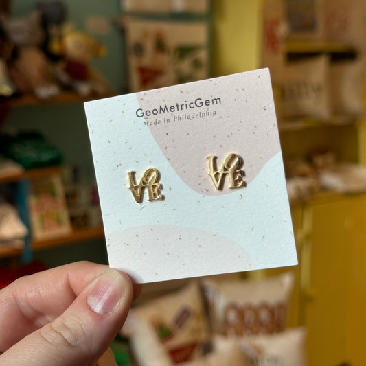 A person holds a card displaying a pair of "LOVE Cutout Earrings." The text on the card reads, "GeoMetricGem - Made in Philadelphia." The gold-plated earrings are designed to be safe for sensitive ears. In the background, various items can be seen on shelves.