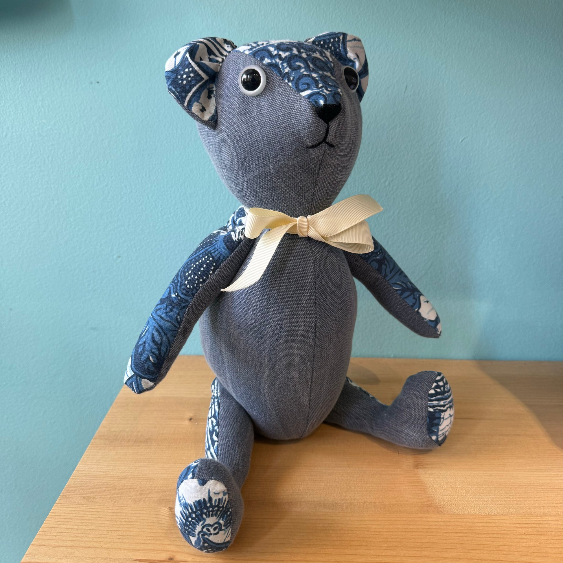 A Stuffed Teddy Bear from Useful & Beautiful Handmade, crafted from blue and white patterned upcycled materials, complete with a cream bow tie and black button eyes, sits on a wooden surface against a light blue background. This charming bear makes an ideal baby gift.