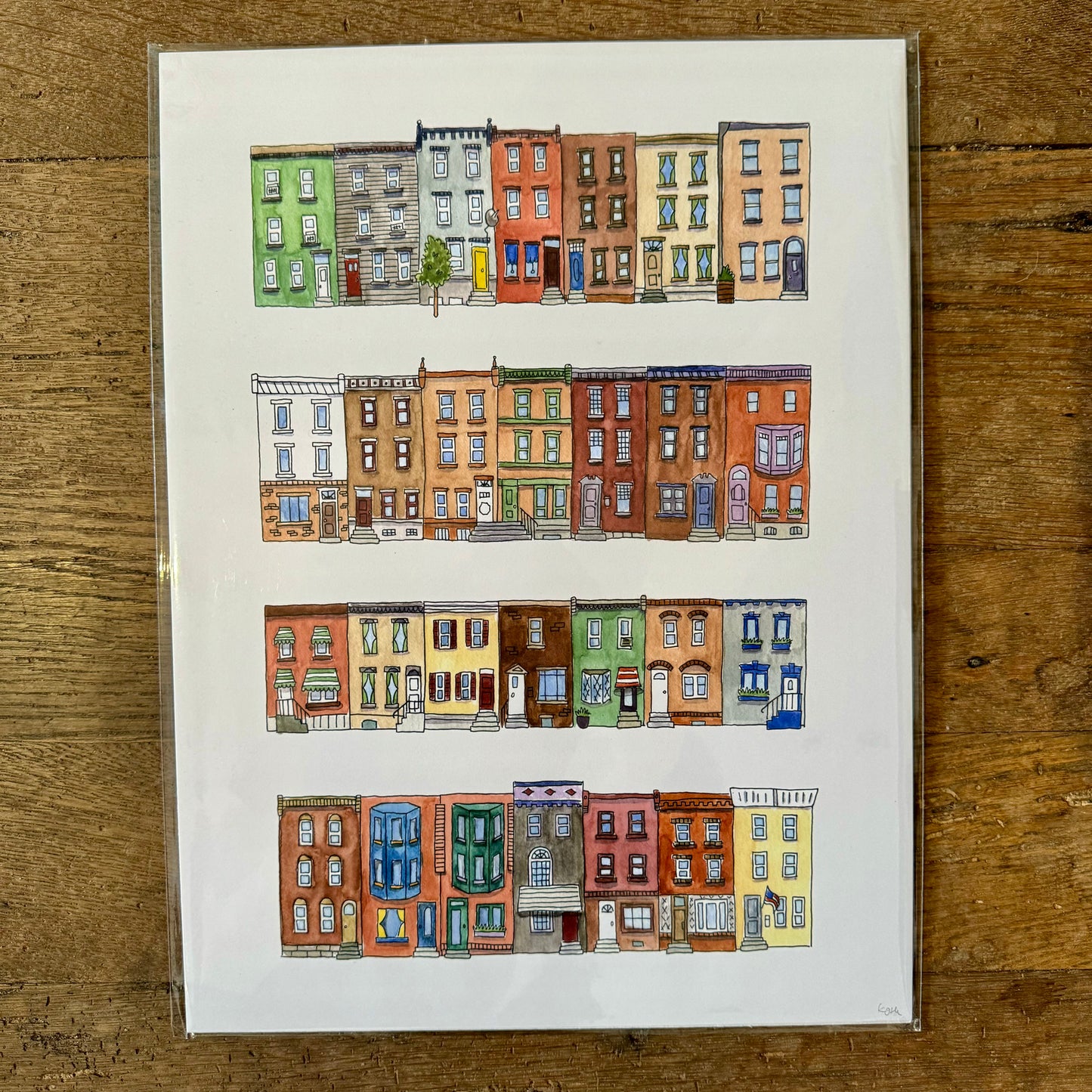 A framed illustration from Kate Otte's Philadelphia Prints collection, showcasing colorful multi-story row houses with varied architectural designs and hues in four horizontal rows, is displayed on a wooden surface.