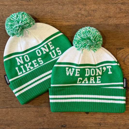 Two South Fellini "No One Likes Us Eagles Beanie II" double-sided knit hats with pom-poms, in green and white, one declaring "NO ONE LIKES US" and the other "WE DON'T CARE," perfect for a Philadelphia Eagles fan reminiscing about the Super Bowl Parade, displayed on a wooden surface.