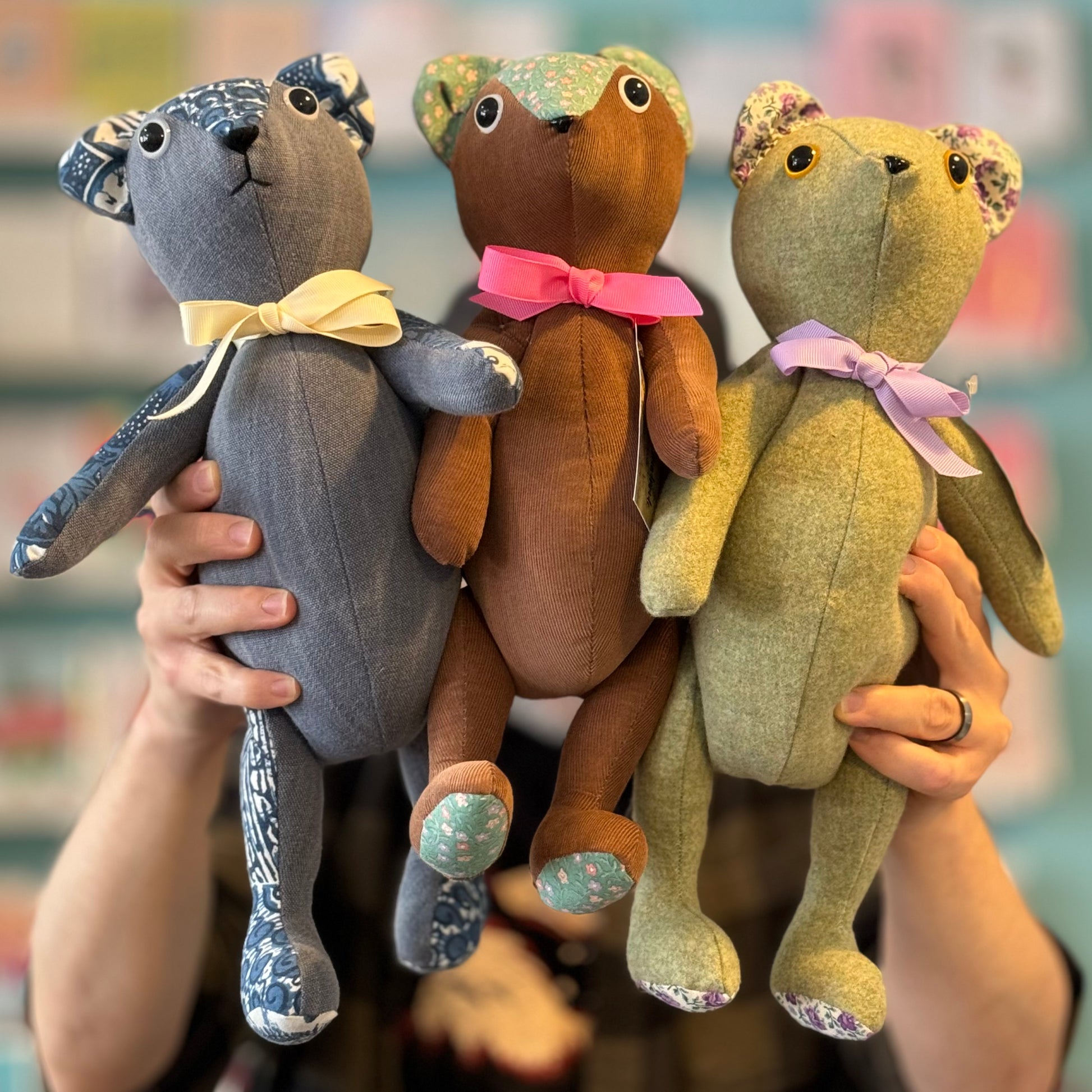 A person holds up three Stuffed Teddy Bears in blue, brown, and green, each featuring patterned fabric patches on their ears and feet made from upcycled materials, along with ribbon bows around their necks. These charming creations by Useful & Beautiful Handmade make the perfect eco-friendly baby gift!